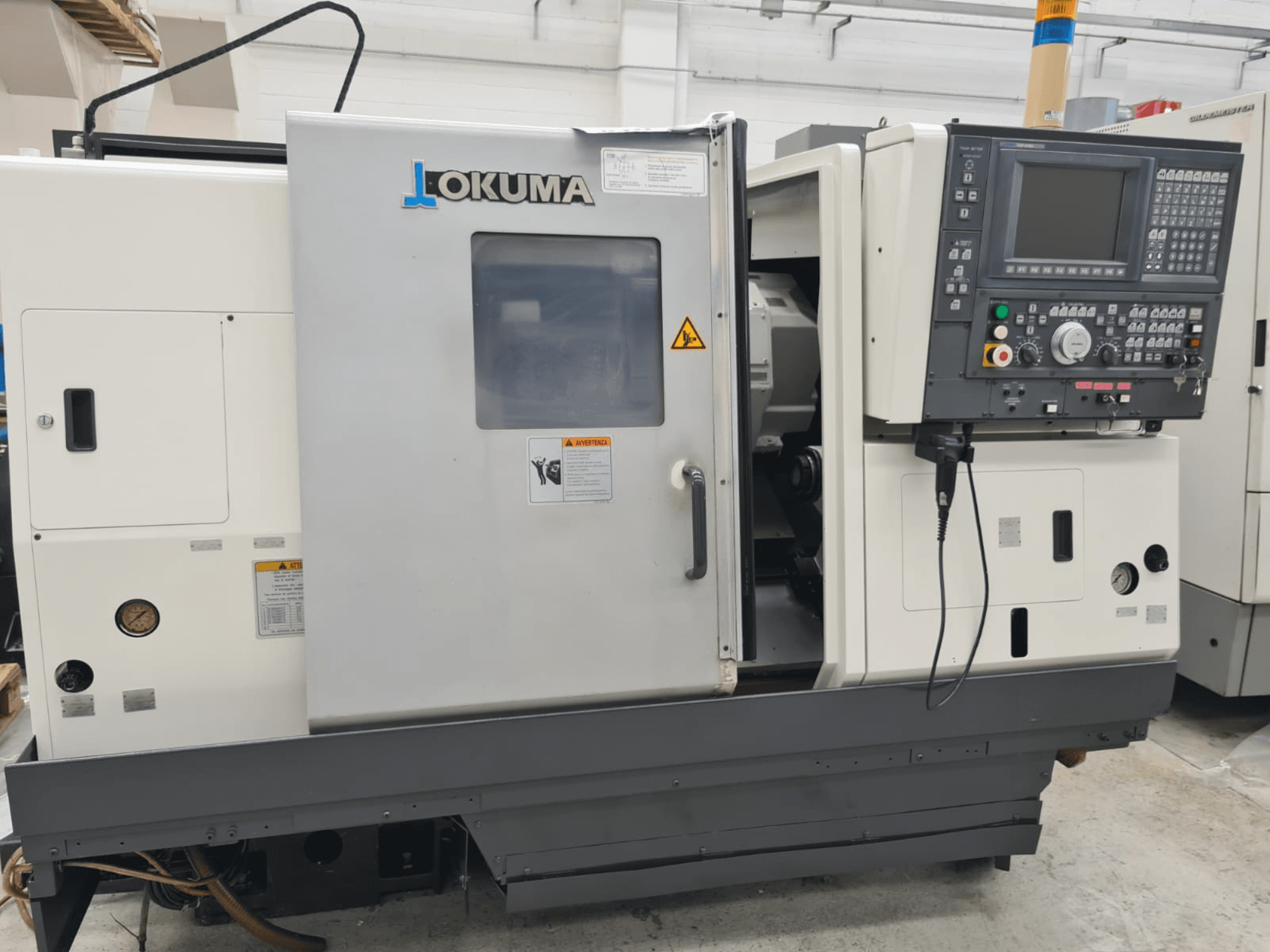 Okuma LB 300M X 500 horizontale draaimachine in zijaanzicht, met het bedieningspaneel, toegang tot de spindel en veiligheidswaarschuwingen.