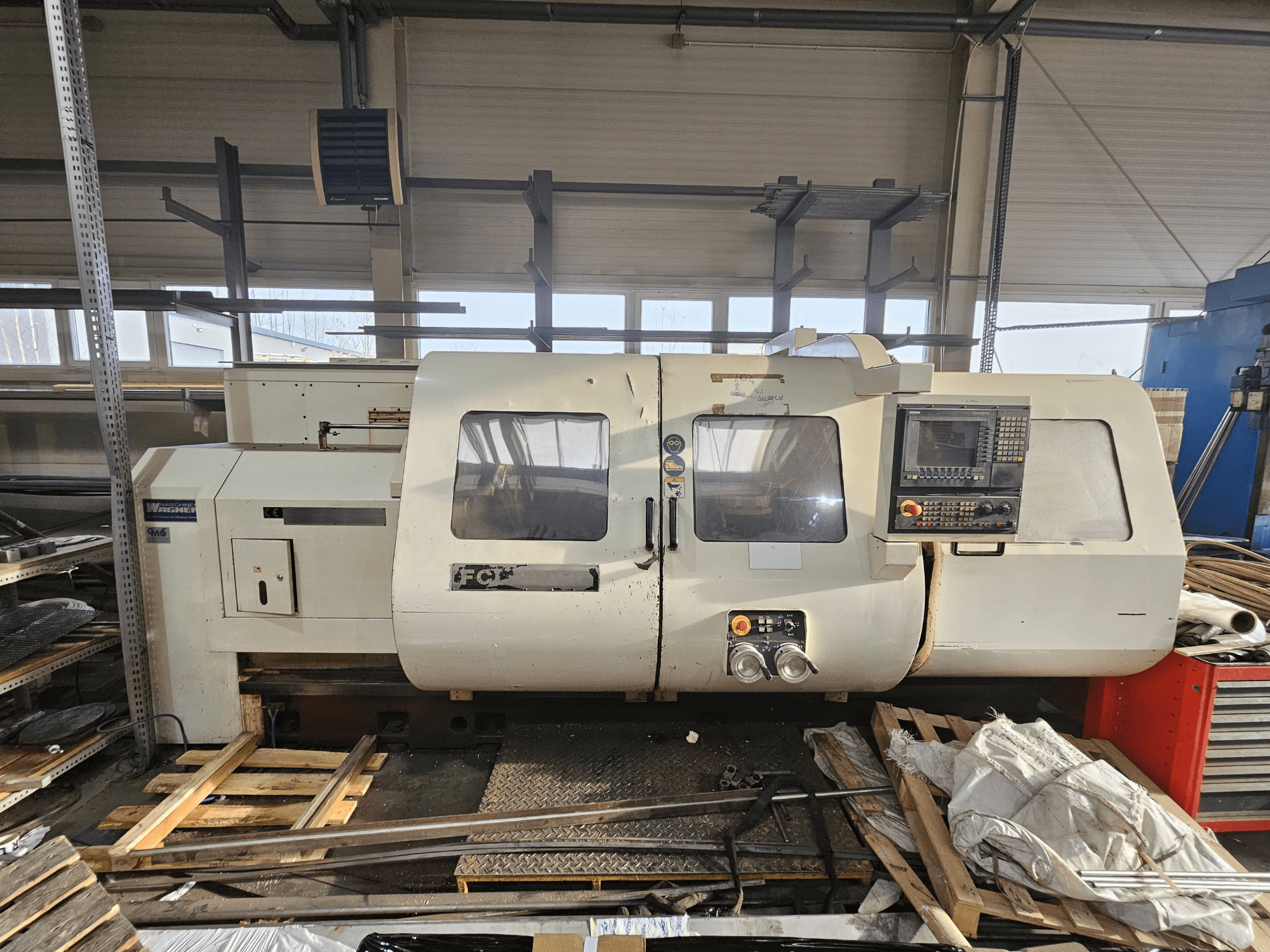 Wabeco CNC-draaibank, zijaanzicht, met bedieningspaneel, ramen en werkgebied; machines op houten pallets in werkplaatsomgeving.