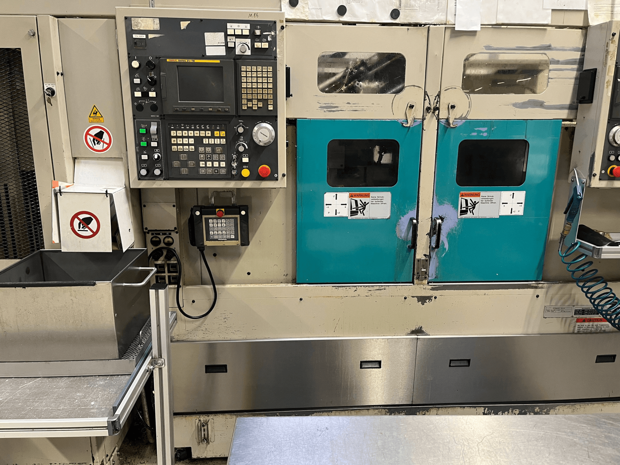 Bedieningspaneel van een productiemachine met groenblauwe deuren, veiligheidswaarschuwingen en een toetsenbord; vooraanzicht, afwerking in roestvrij staal.