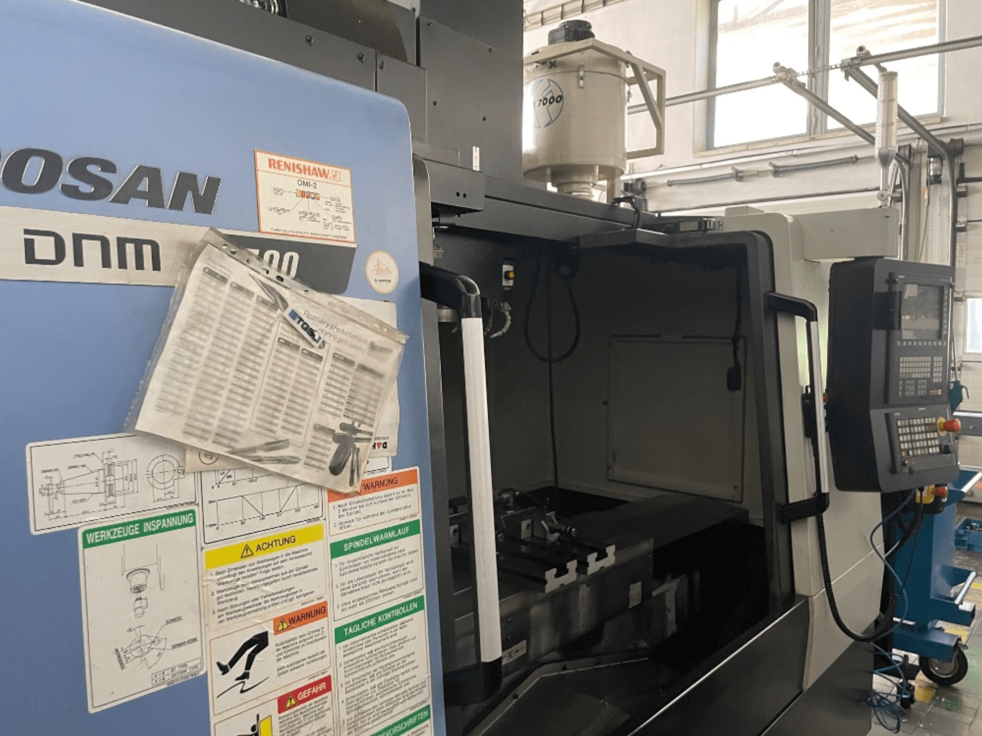 DOOSAN DNM 5700 verticaal bewerkingscentrum, vooraanzicht, toont bedieningspaneel, veiligheidslabels en machinedetails.