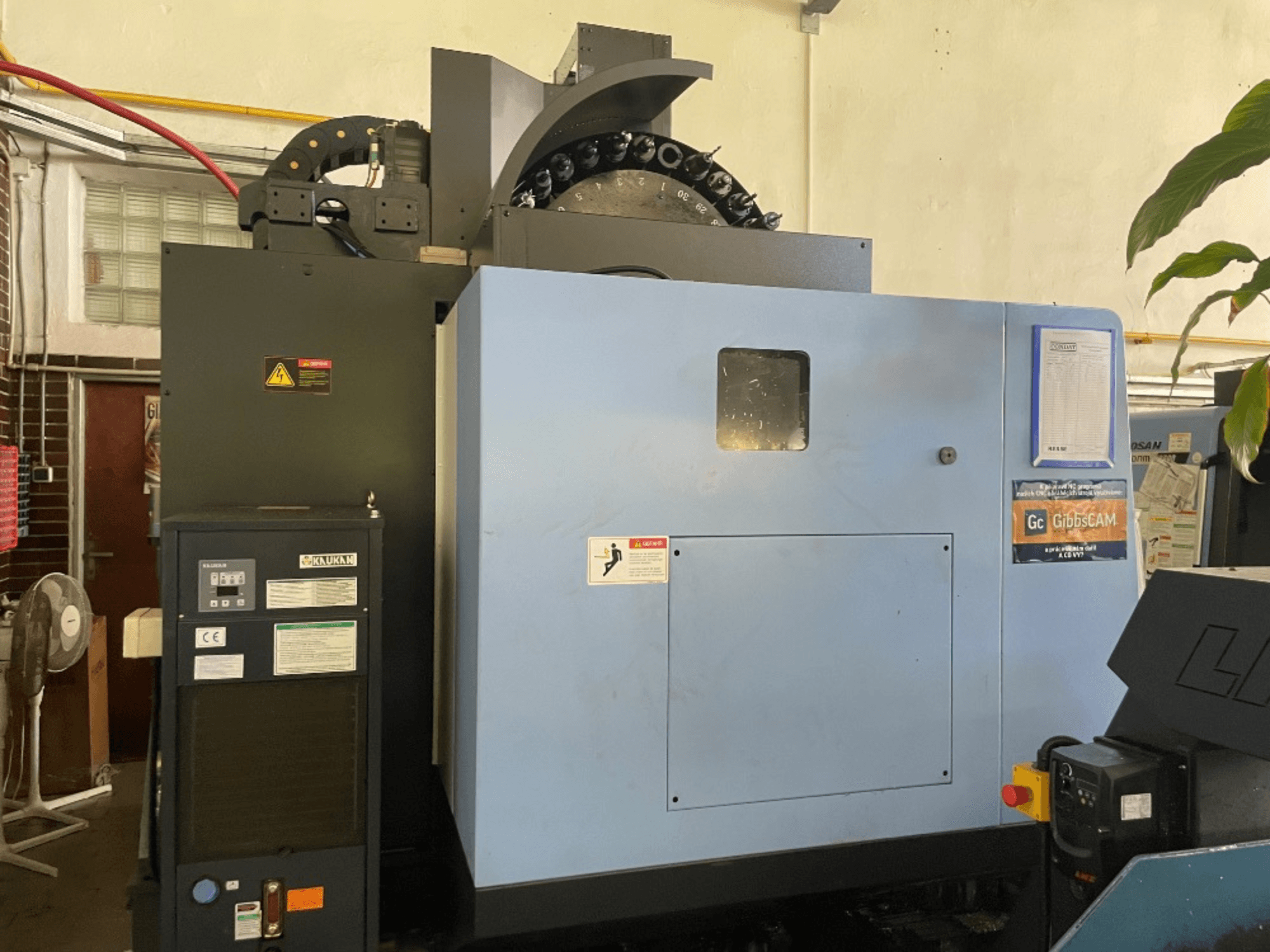 Vooraanzicht van een DOOSAN DNM 5700 verticaal bewerkingscentrum, met het bedieningspaneel, veiligheidslabels en machinekenmerken.