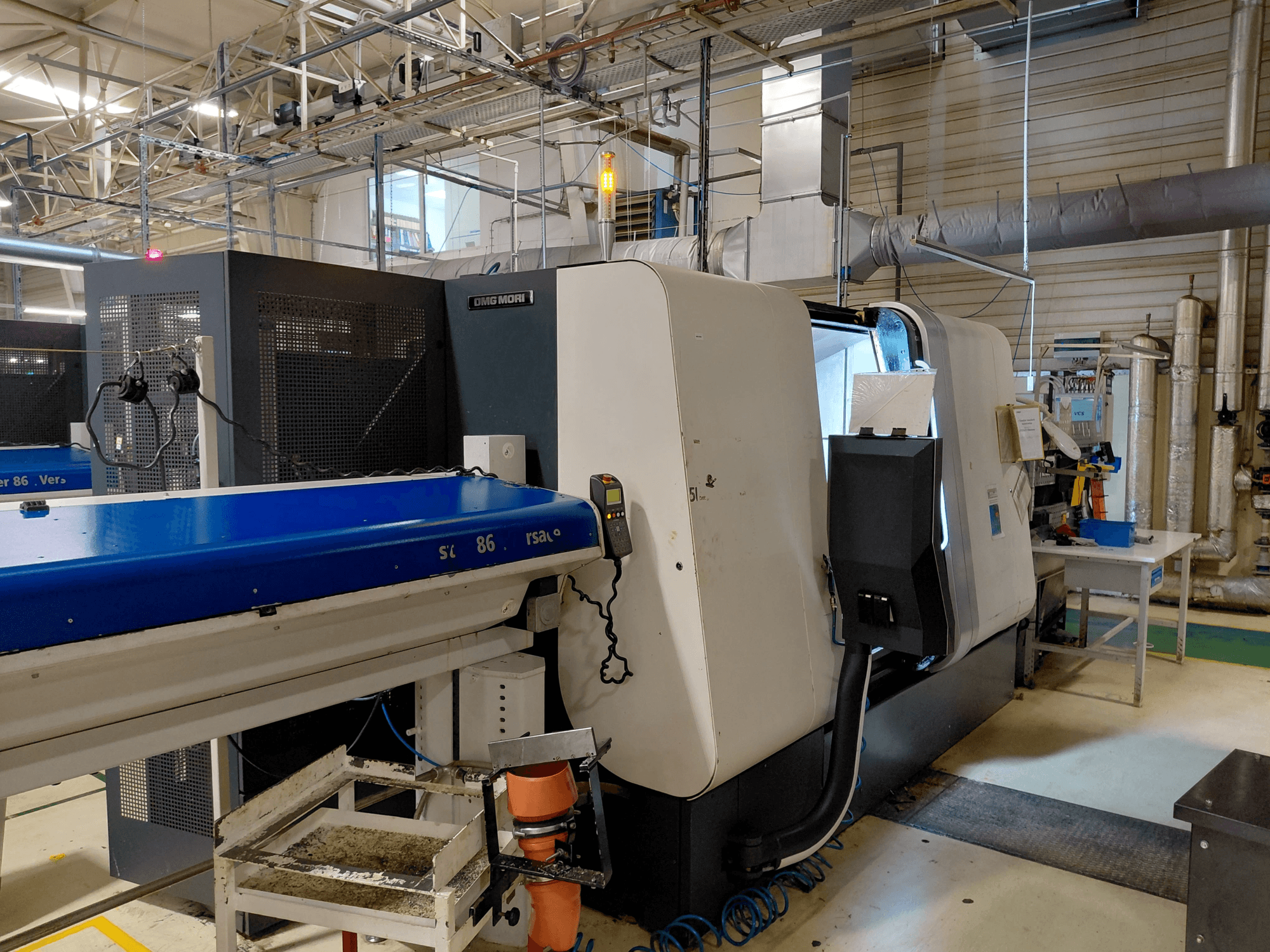 DMG MORI Sprint 65 lineaire meerspillige draaimachine, zijaanzicht met bedieningspaneel en transportband zichtbaar in een werkplaats.