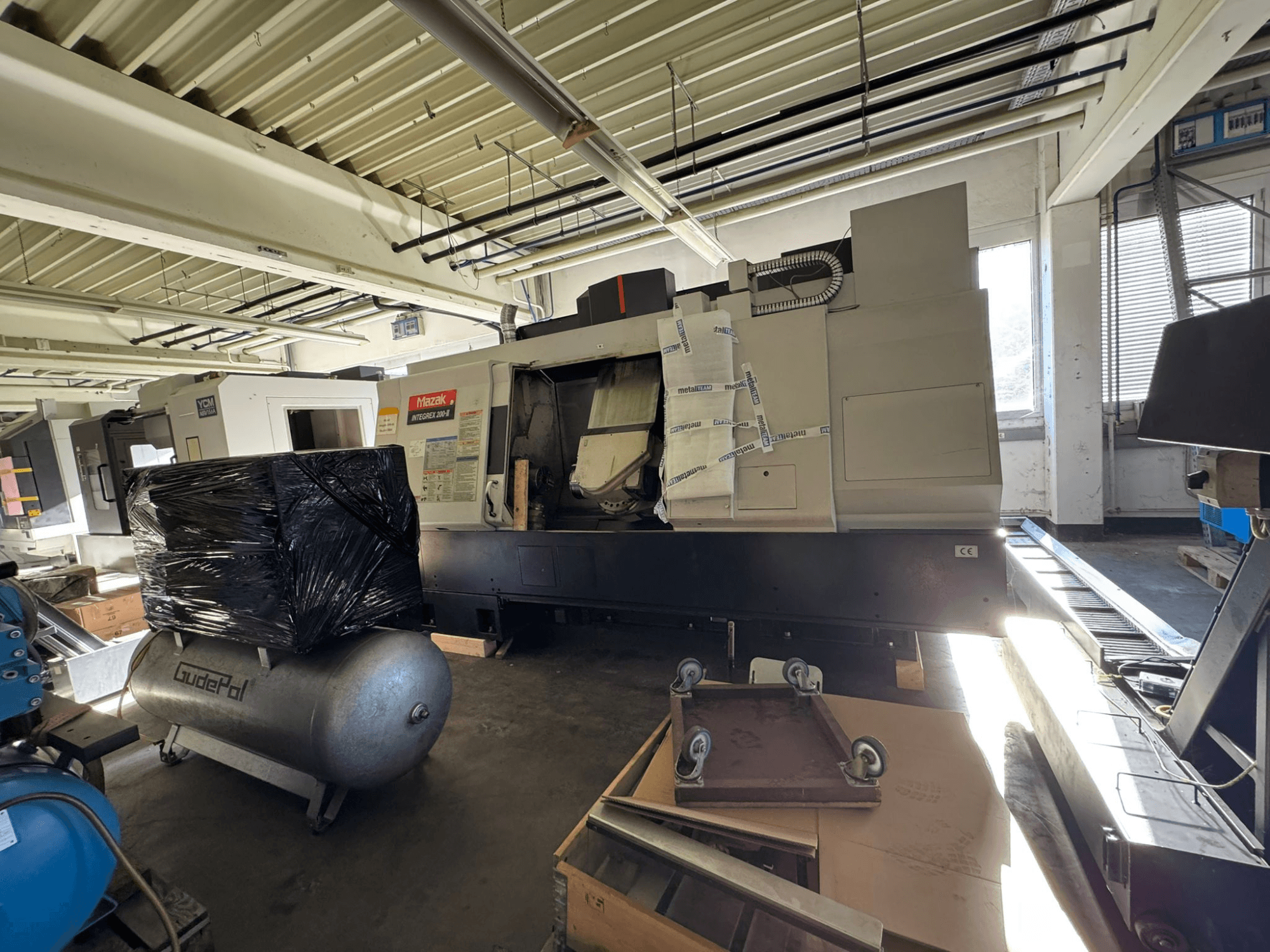 Vooraanzicht van het Mazak INTEGREX 200-3 frees-draaicentrum in een werkplaats, gedeeltelijk aan het zicht onttrokken door verpakkingsmateriaal en apparatuur in de buurt.