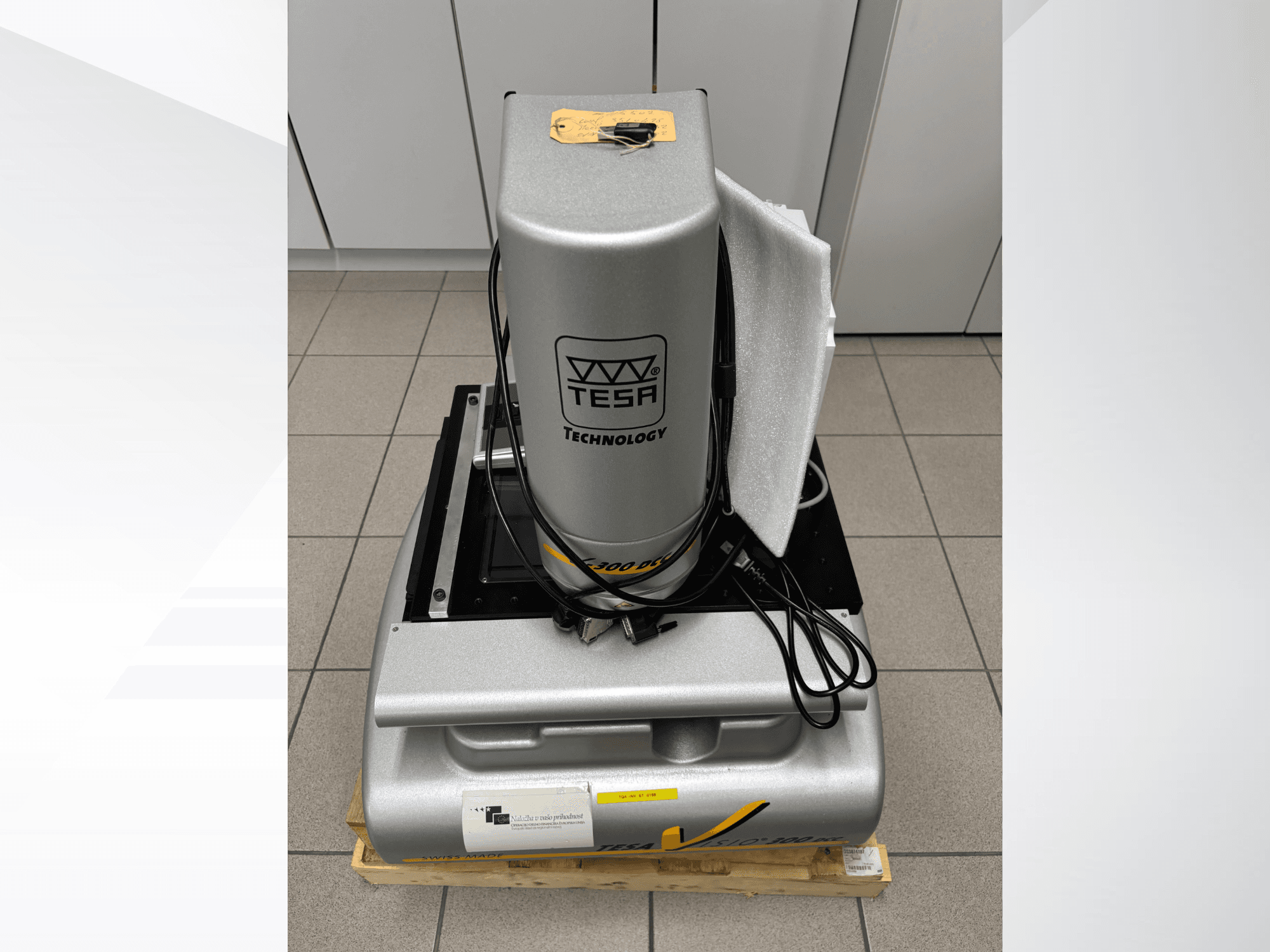 TESA Visio 300 DCC optische meetmachine in vooraanzicht met kabels, op een houten pallet, en beschermende verpakking zichtbaar.