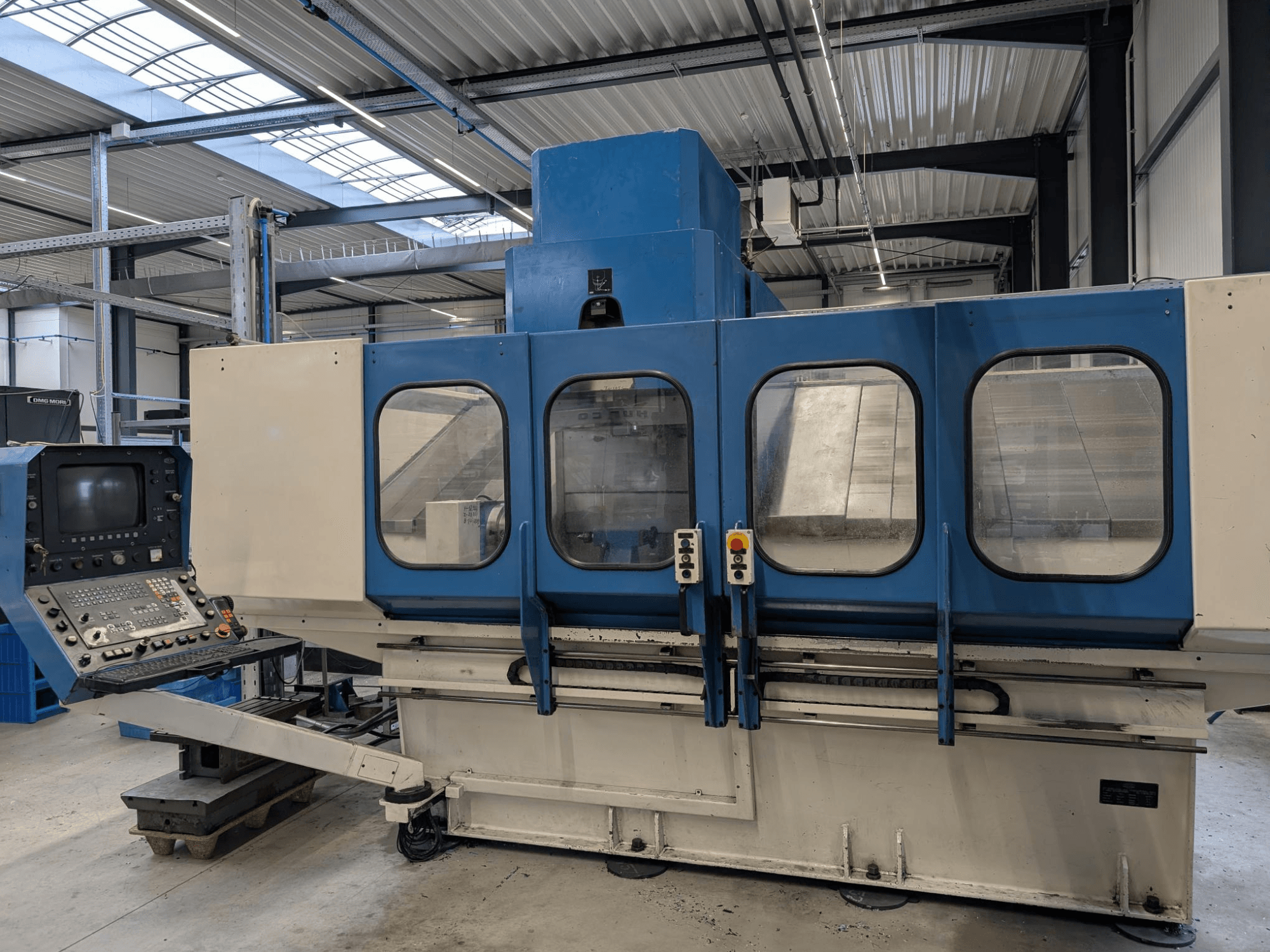 Blauwe industriële CNC machine met bedieningspaneel, vooraanzicht. Grote glazen vensters, zichtbare machineonderdelen en knoppen.