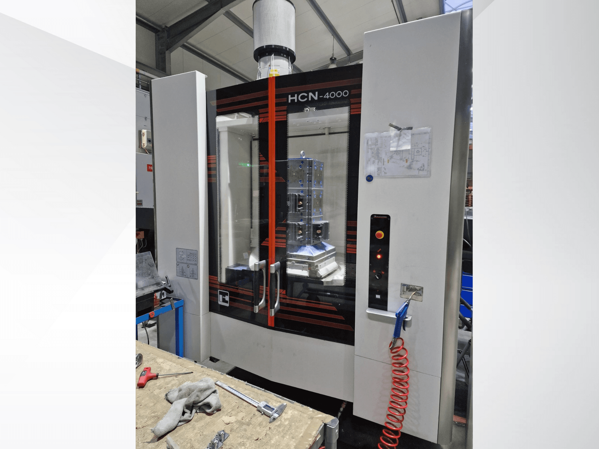 HCN-4000 CNC machine in vooraanzicht, met een glazen paneel, bedieningspaneel met knoppen en een bevestigde blauwdruk.