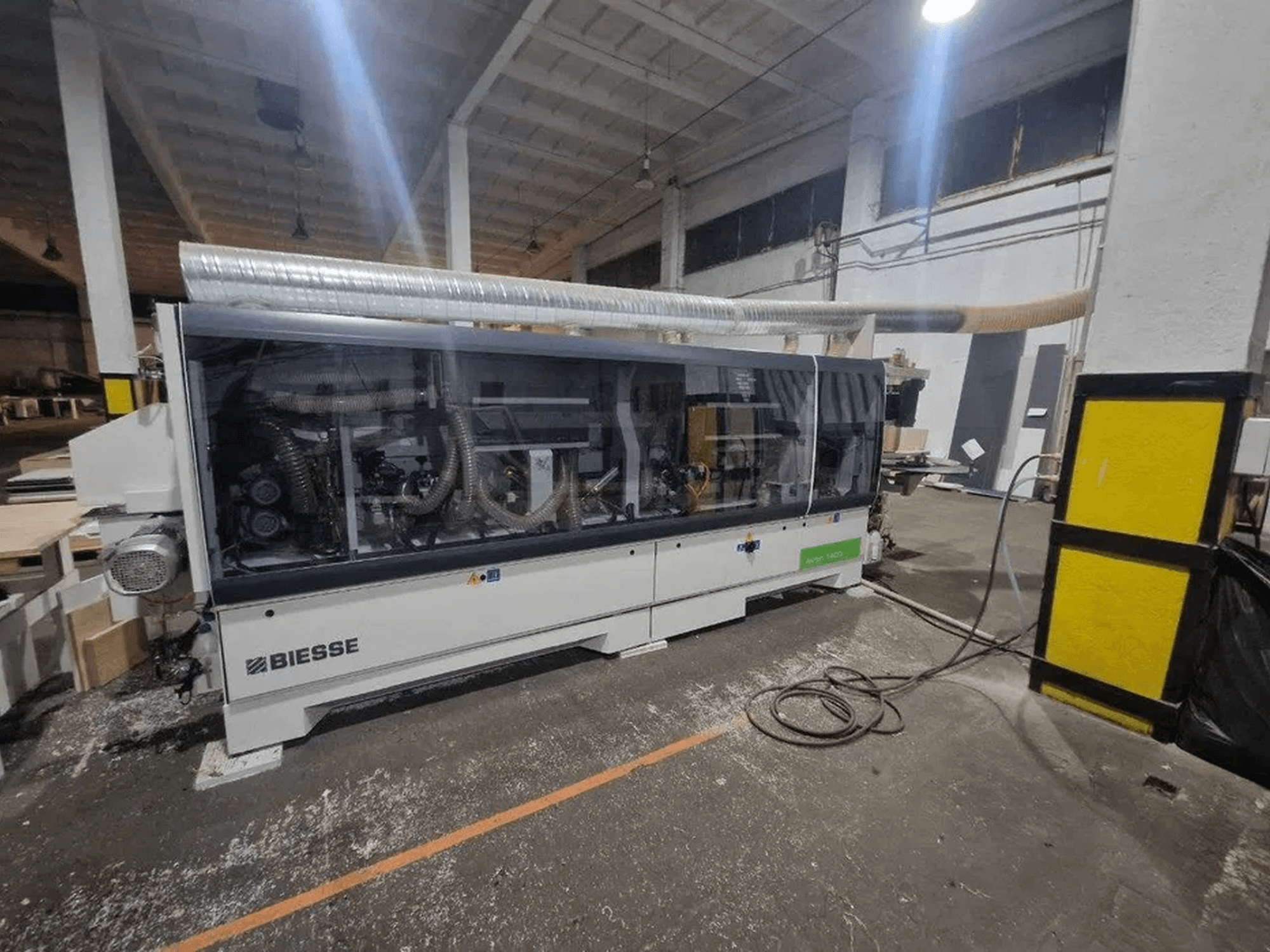Een BIESSE Akron 1440 A-kantenlijmmachine in een werkplaats, met een doorzichtige afdekking en zichtbare machineonderdelen.