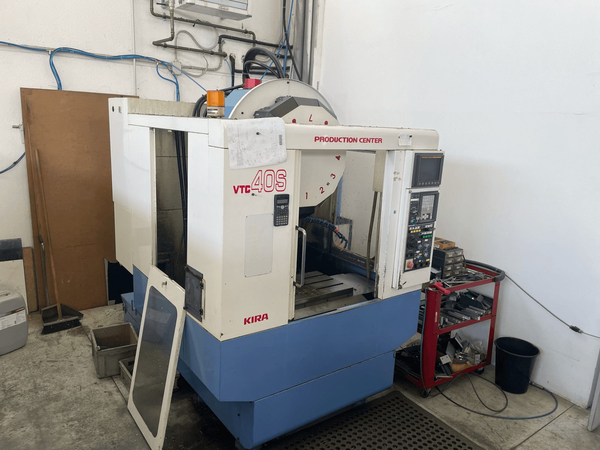 KIRA VTC 40S bewerkingscentrum in zijaanzicht, met bedieningspaneel, open deur en gereedschappen op een nabijgelegen opslagwagentje.