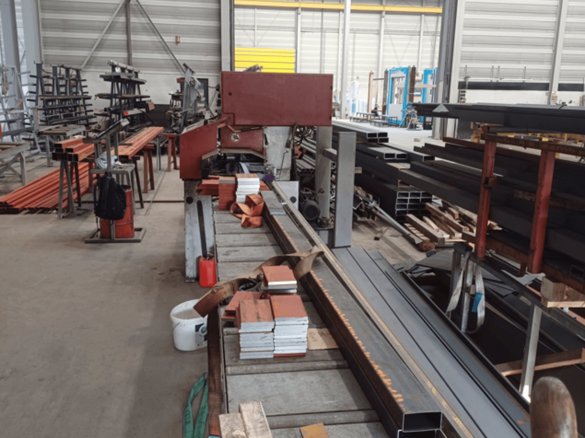 Productiewerkplaats met verschillende metalen profielen, met een rode zaagmachine op de voorgrond en gestapelde materialen.