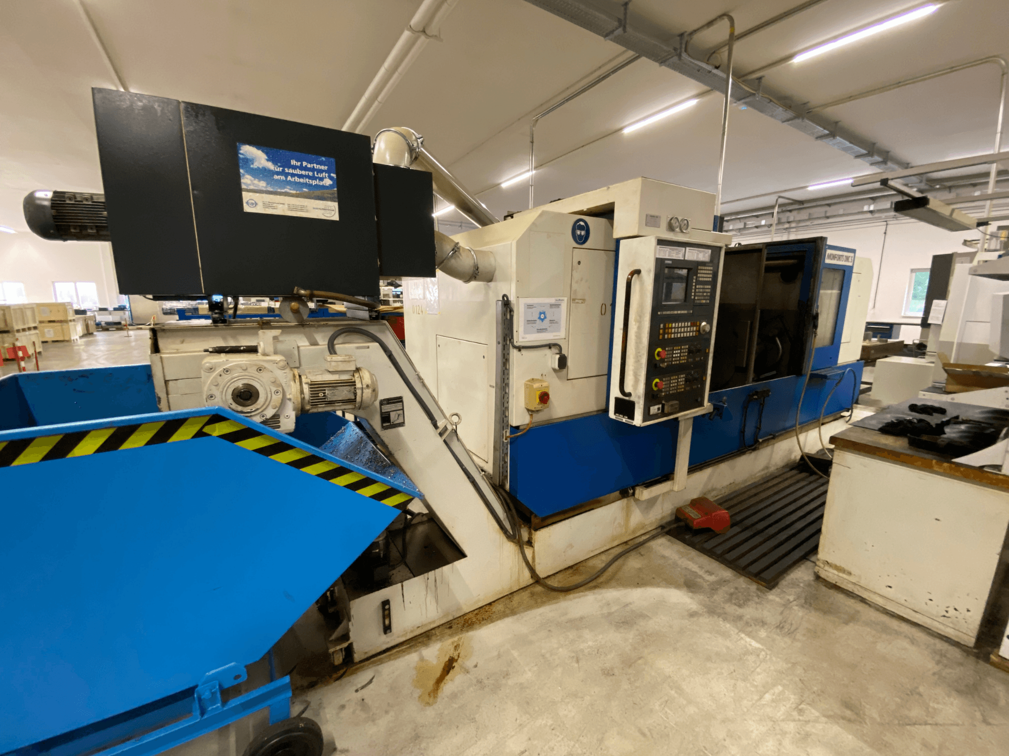 Monforts DNC 5 draai-freescentrum, zijaanzicht met bedieningspaneel, blauwe spanenbak en machineonderdelen in een werkplaatsomgeving.