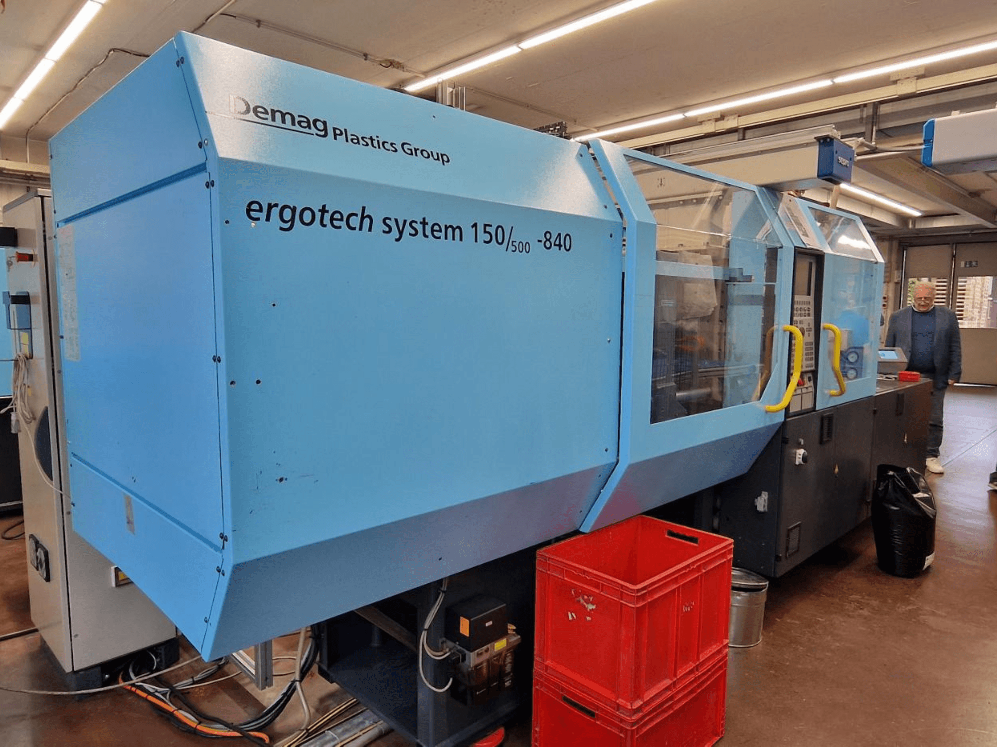 Demag Ergotech System 150/500-840 spuitgietmachine in blauw, zijaanzicht, met kratten en operator op de achtergrond.