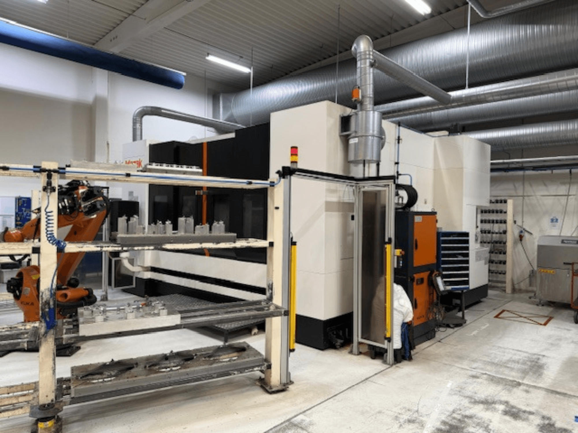 Industriële CNC machine met robotarm en veiligheidshekken, met vooraanzicht van de opstelling in een luchtige werkruimte.