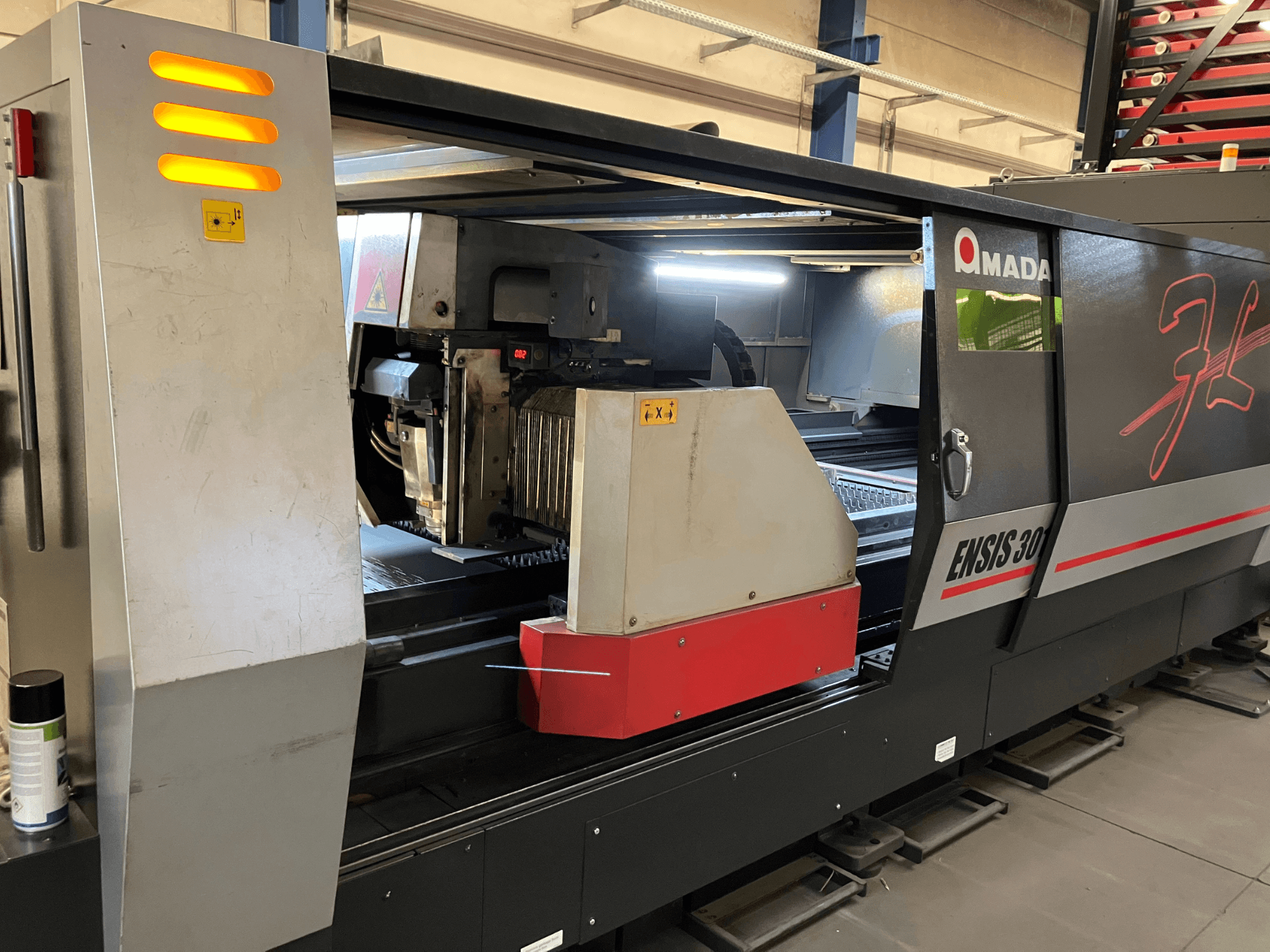 AMADA ENSIS 3015 AJ fiberlasersnijmachine, vooraanzicht, met snijkop en bedieningselementen in een metalen frame.