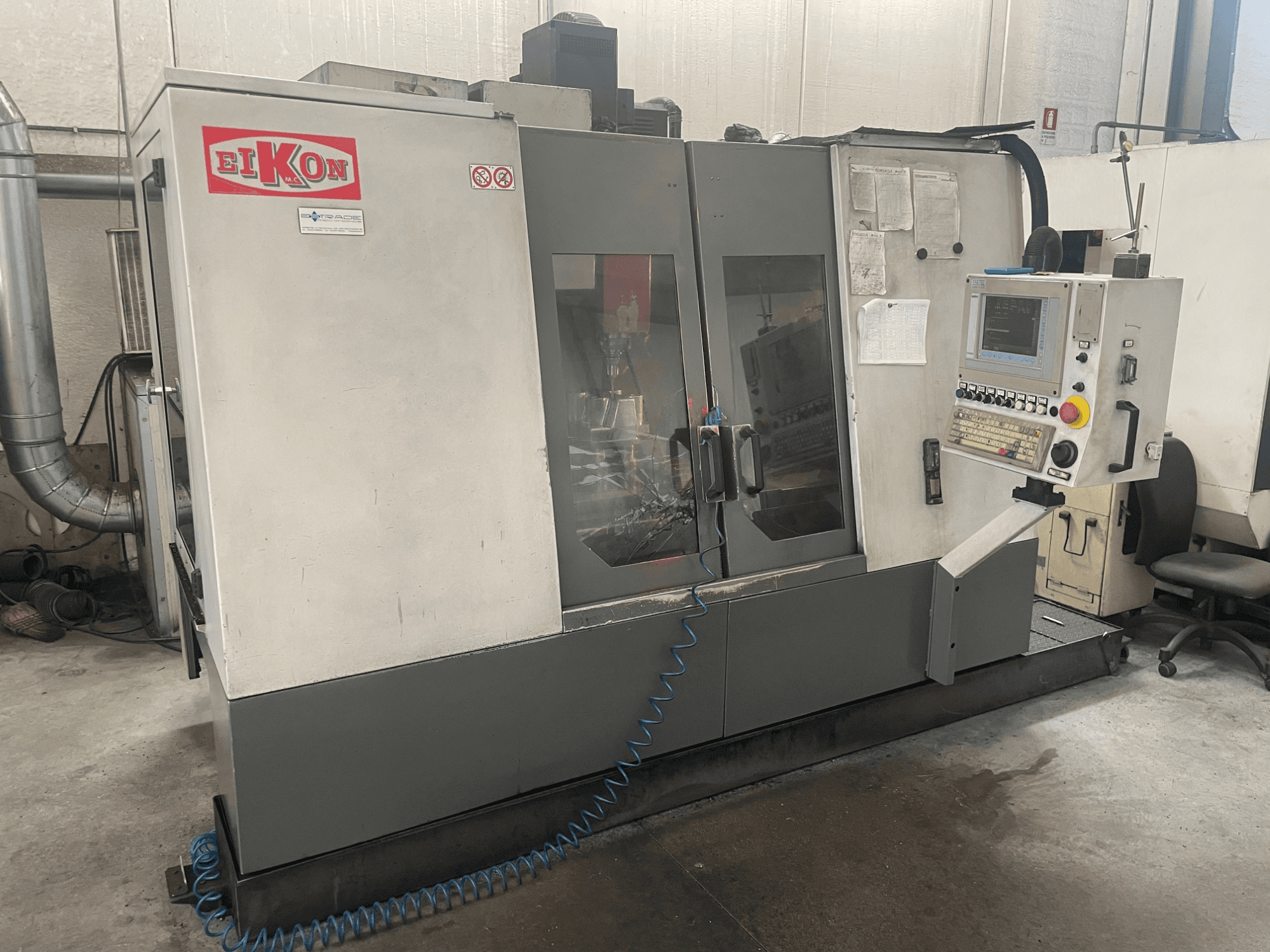 EIKON CNC machine gezien vanaf de voorkant, met een bedieningspaneel, grote glazen deuren en een blauwe luchtslang op de vloer.