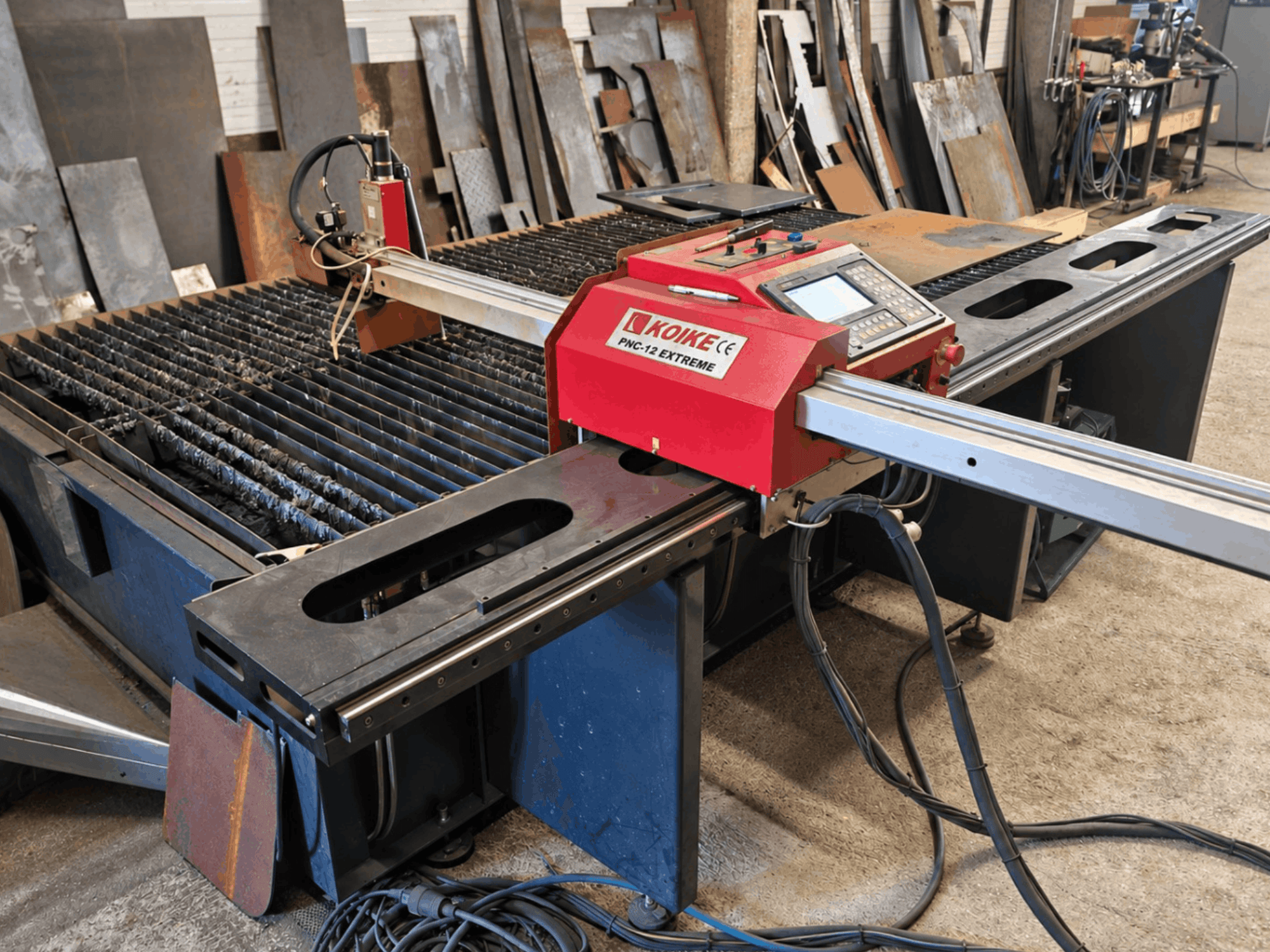 Vooraanzicht van de KOIKE PNC-12 Extreme plasmasnijmachine met metalen snijtafel, bedieningspaneel en kabels.