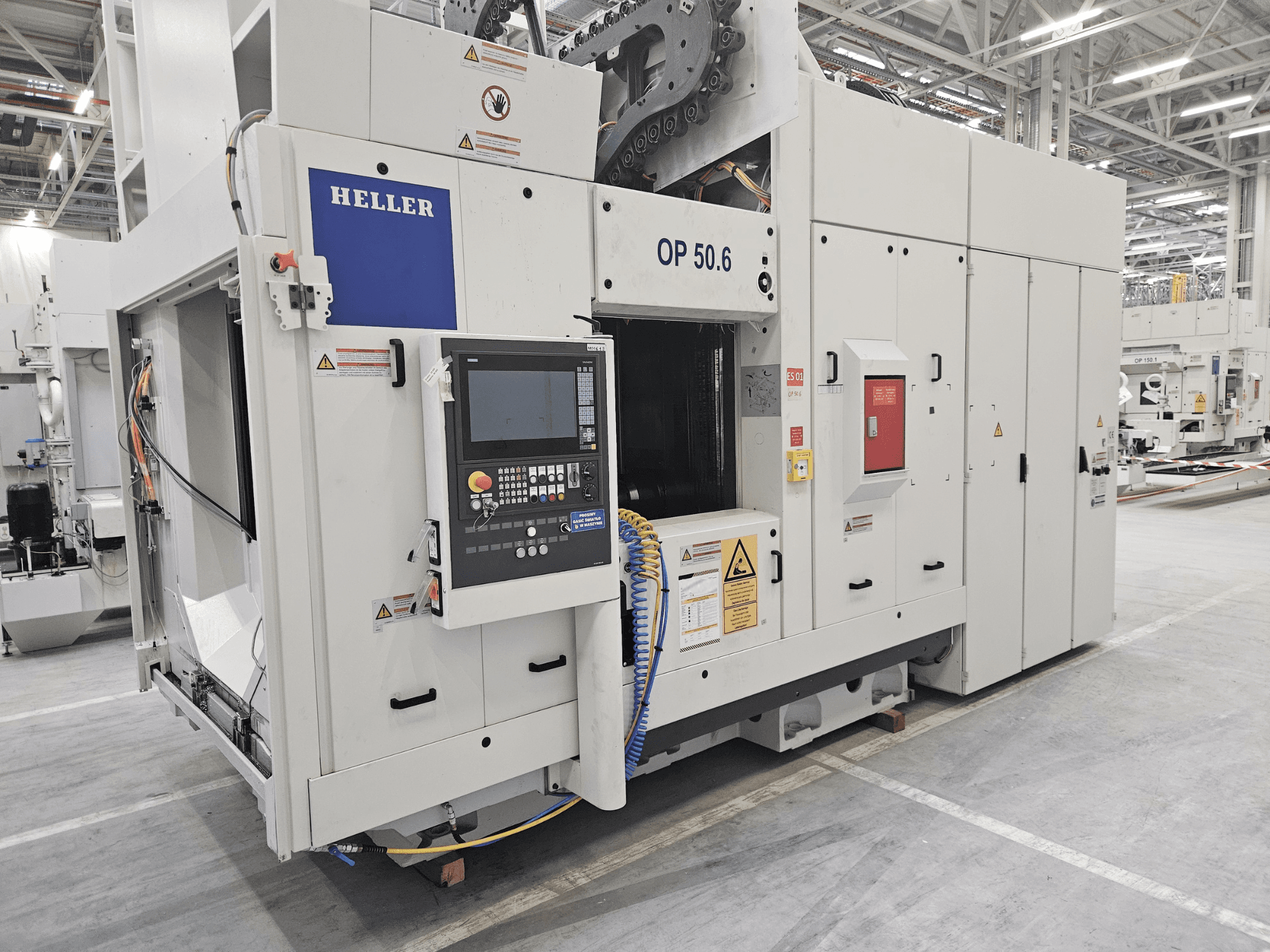 Heller OP 50.6 bewerkingscentrum, vooraanzicht, met een bedieningspaneel, waarschuwingslabels en slangen zichtbaar in een fabrieksomgeving.