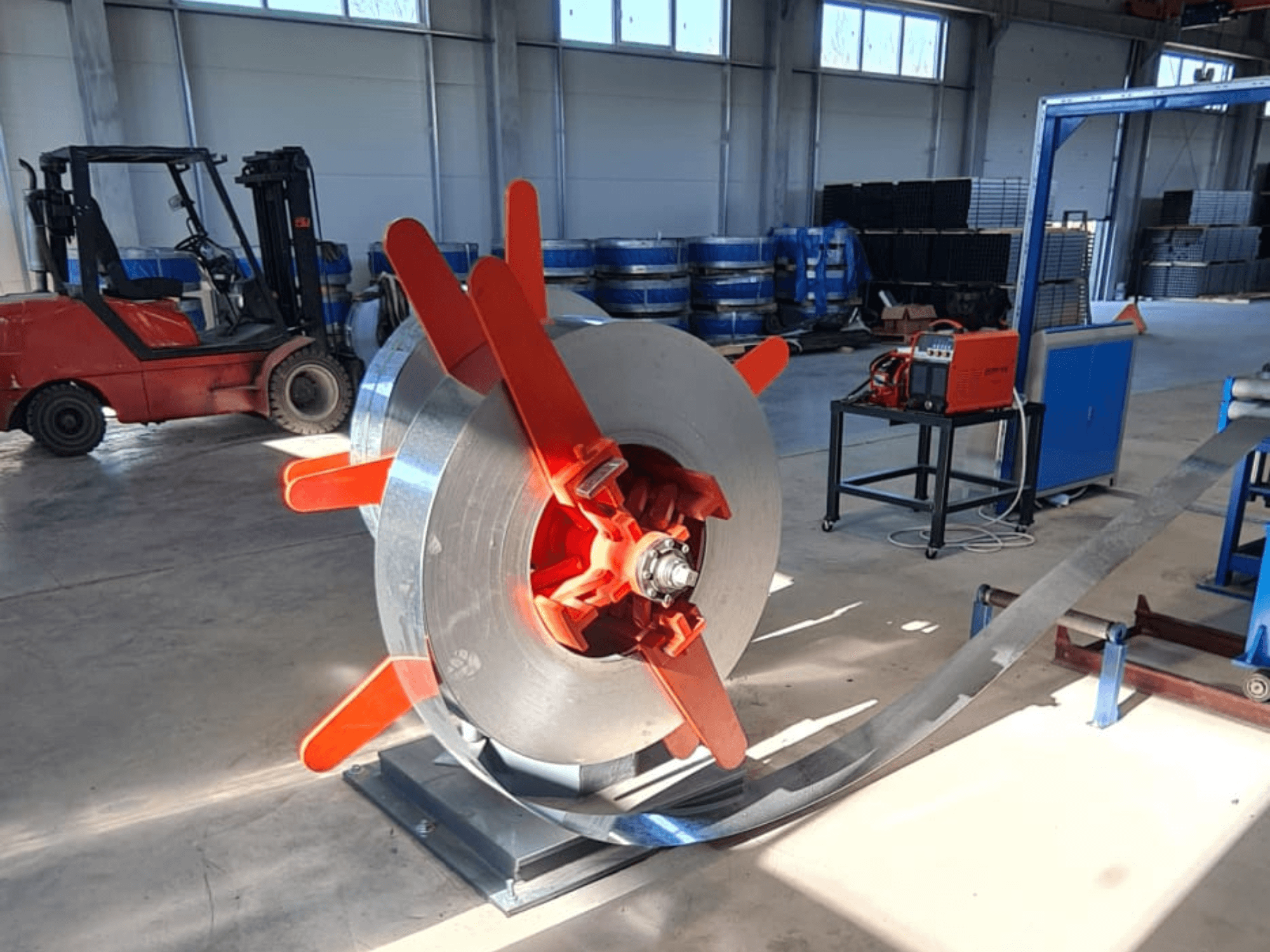 Metalen spoel met rode handgrepen op een standaard, gezien vanuit een hoek. Industriële achtergrond met machines en een vorkheftruck.