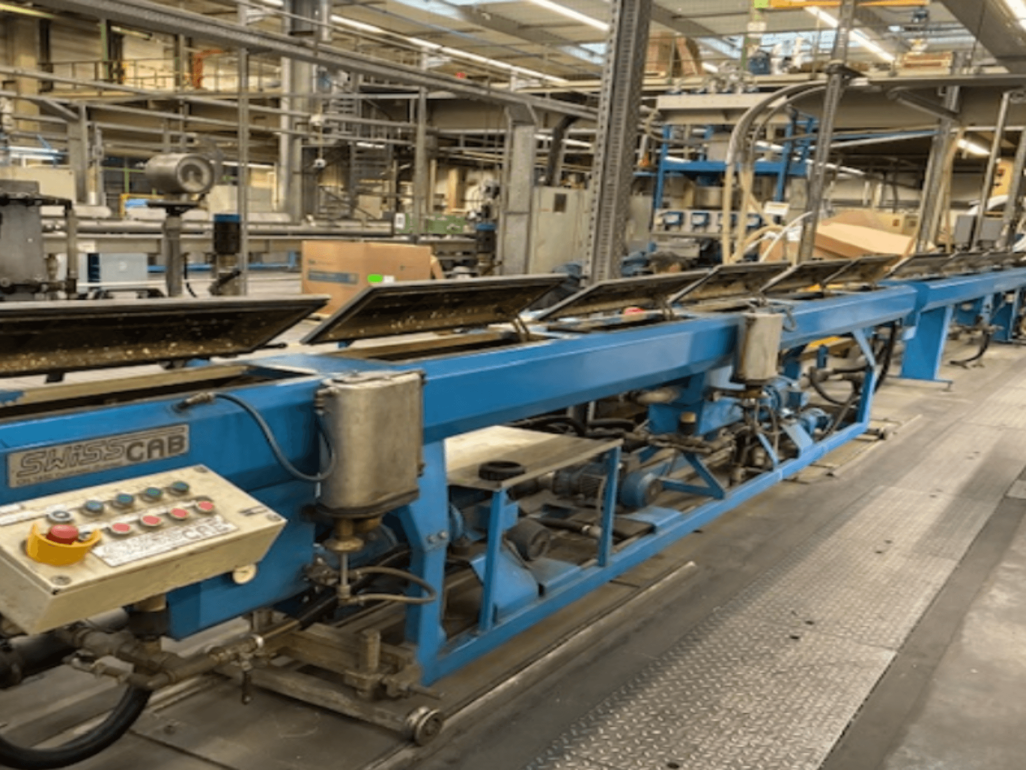 Blauwe SwissCab industriële machine in een fabrieksomgeving, met bedieningsknoppen en een transportsysteem, zijaanzicht.