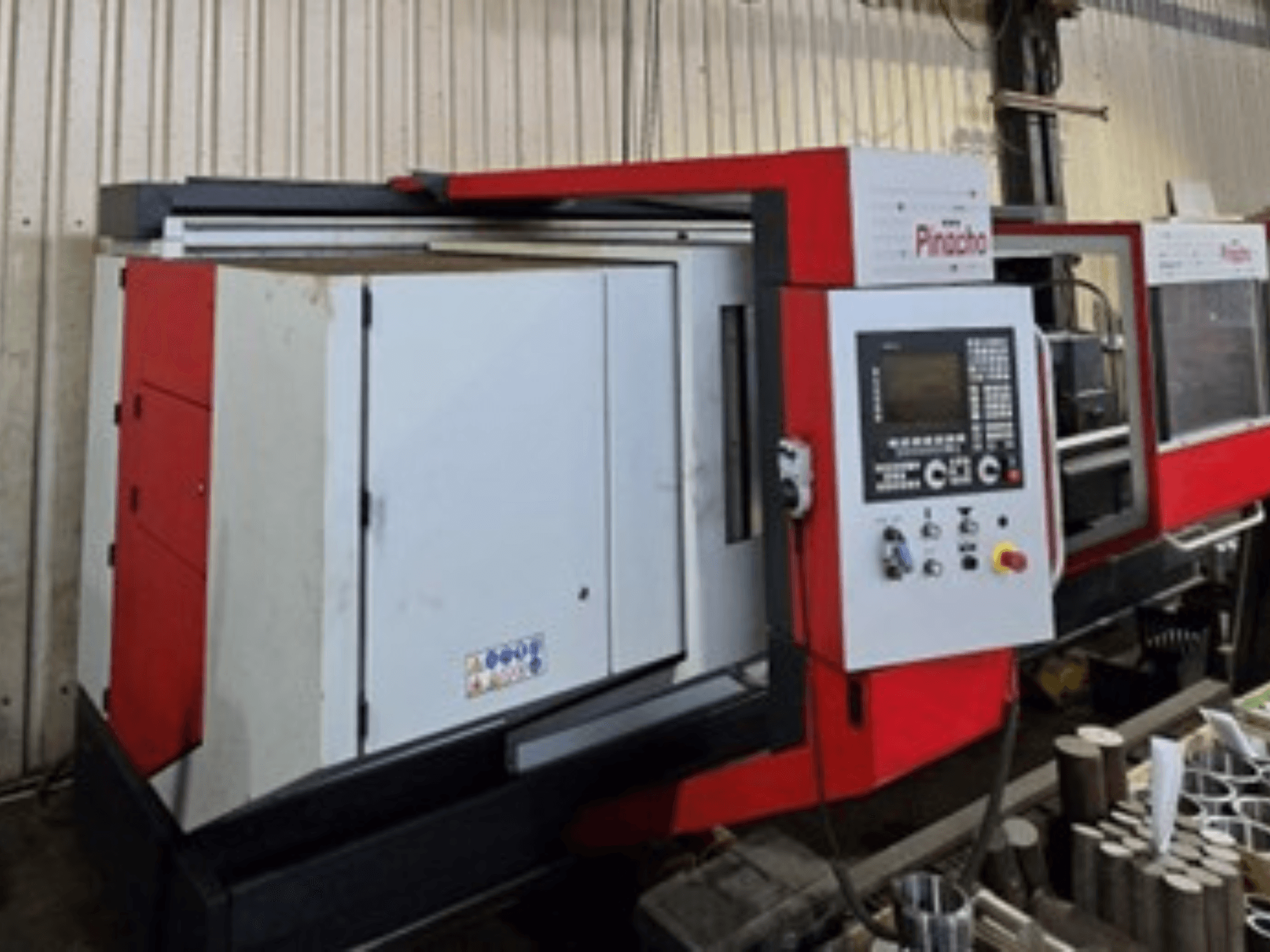 Pinacho CNC machine in rood en wit, zijaanzicht, met bedieningspaneel en materialen op de vloer, wat de operationele setup toont.