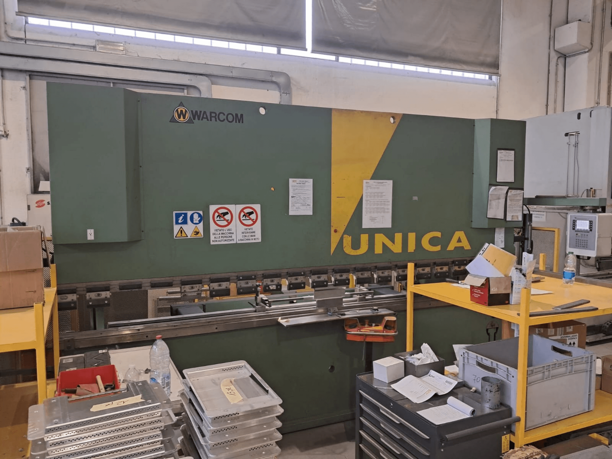 Hydraulische pers Warcom UNICA 100-40, vooraanzicht, groene behuizing met gele accenten, veiligheidsborden zichtbaar, gereedschap en materialen op de tafel.