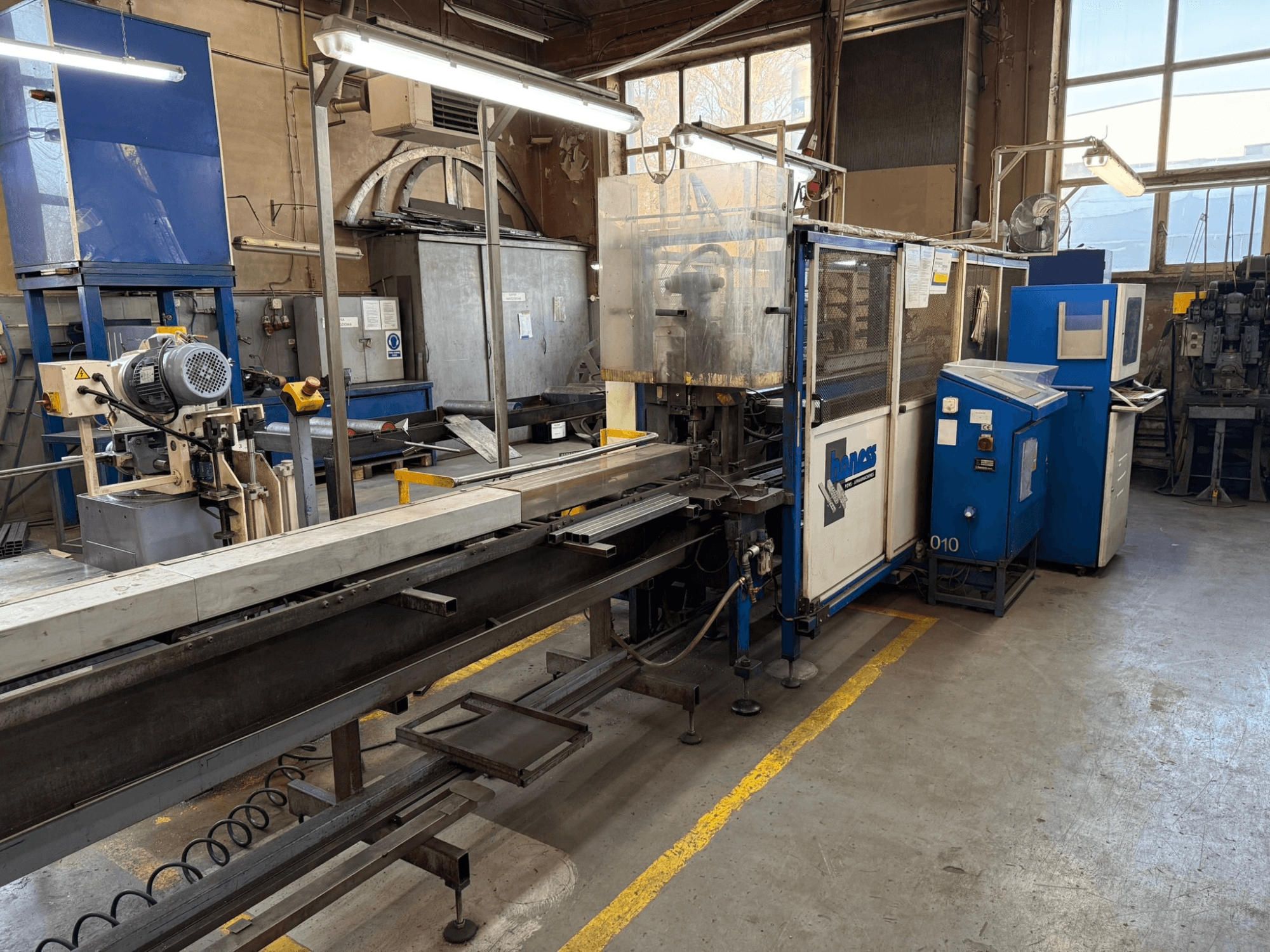 Industriële werkplaats met een blauwe Haweas machine, zijaanzicht met transportband en apparatuur in een grote, goed verlichte ruimte.