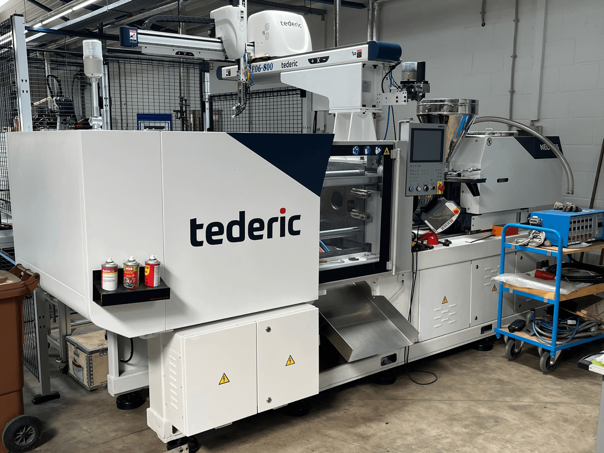Hydraulische spuitgietmachine TEDERIC NEO.E55/e110h in een werkplaats, zijaanzicht met bedieningspaneel en aangrenzende gereedschappen.