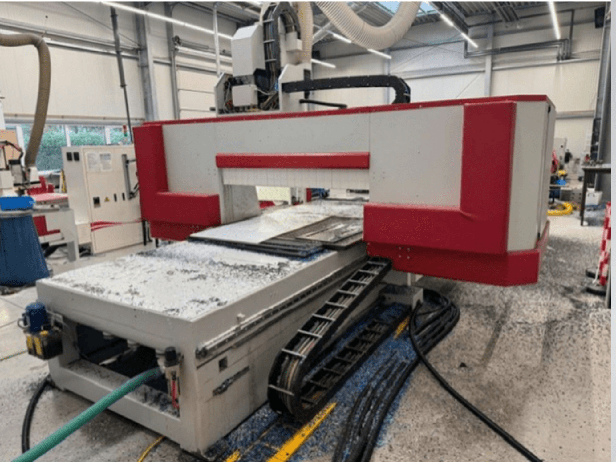 Rood met grijze CNC machine in een werkplaats, zijaanzicht met materiaaltafel, koelvloeistofslang en zaagsel op de vloer.