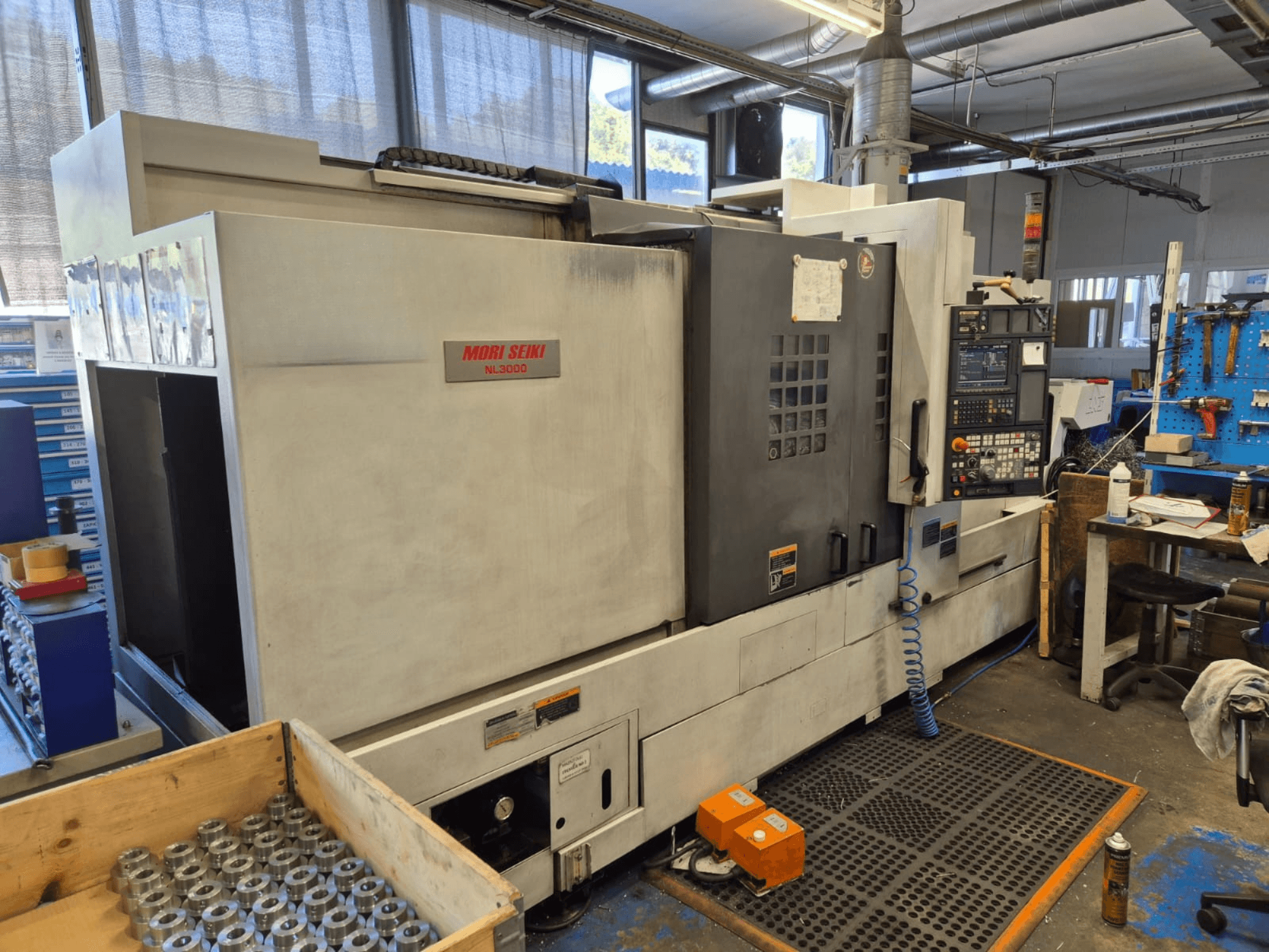 Mori Seiki NL3000 CNC draaibank in een werkplaats, zijaanzicht met bedieningspaneel, gereedschappen en metalen onderdelen in een houten kist.