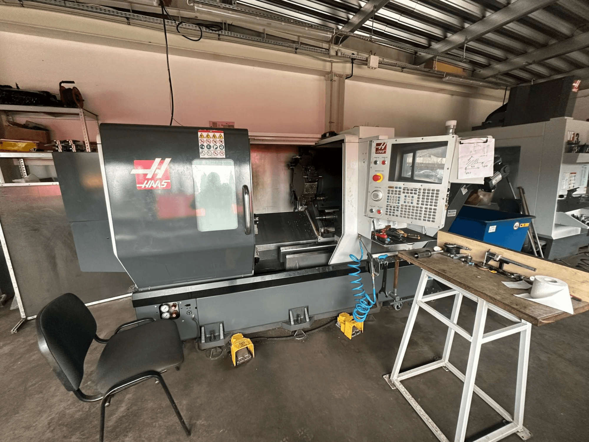 Haas CNC-draaibank (model ST-10) in een werkplaats, zijaanzicht met bedieningselementen, gereedschap op een werkbank en een stoel in de buurt.