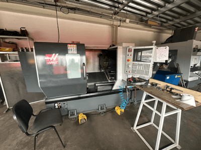 Haas CNC-draaibank (model ST-10) in een werkplaats, zijaanzicht met bedieningselementen, gereedschap op een werkbank en een stoel in de buurt.