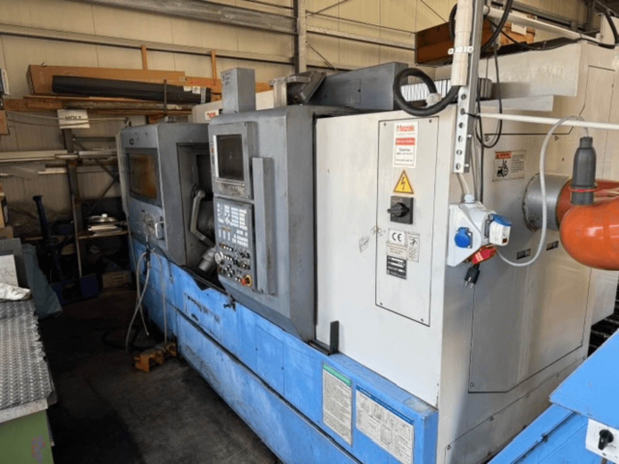 Mazak SQT-30M * 1000 draai-freescentrum, zijaanzicht met bedieningspaneel, blauwe voet en omringende apparatuur in de werkplaats.