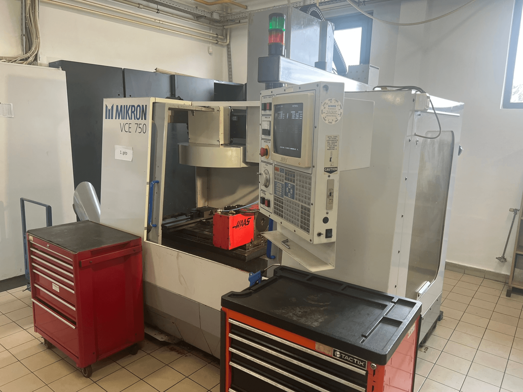 Alt-tekst: "MIKRON VCE 750 verticaal bewerkingscentrum, vooraanzicht met bedieningspaneel en rode gereedschapskisten, in een ruime werkplaats."