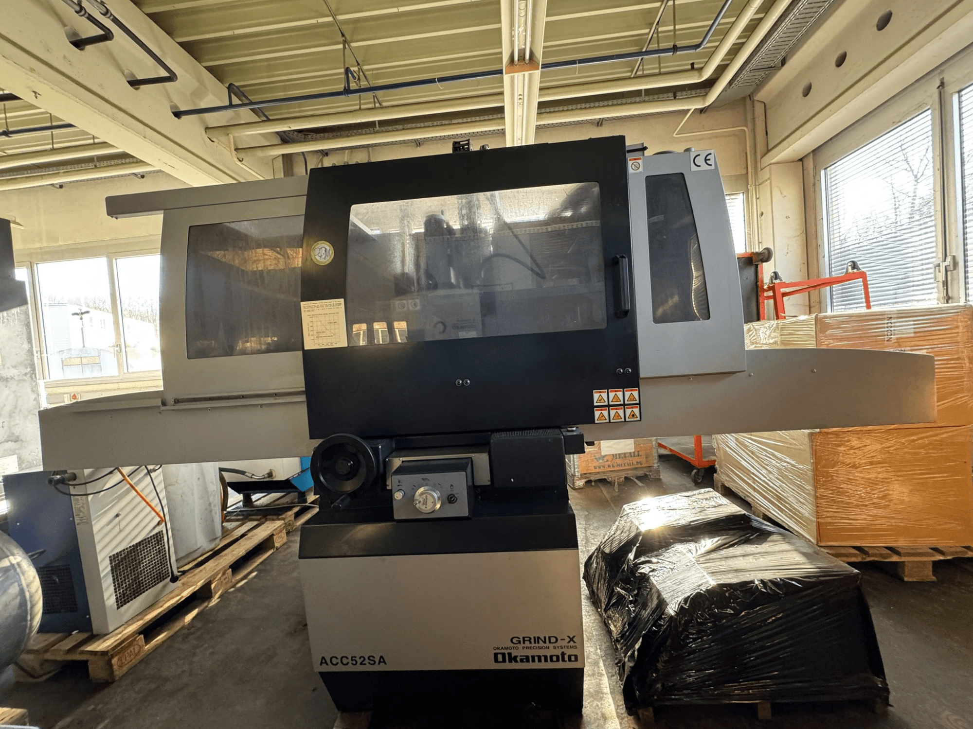 Okamoto ACC 52 SA-vlakkenslijpmachine in zijaanzicht, met bedieningselementen en labels, omringd door pallets en dozen.