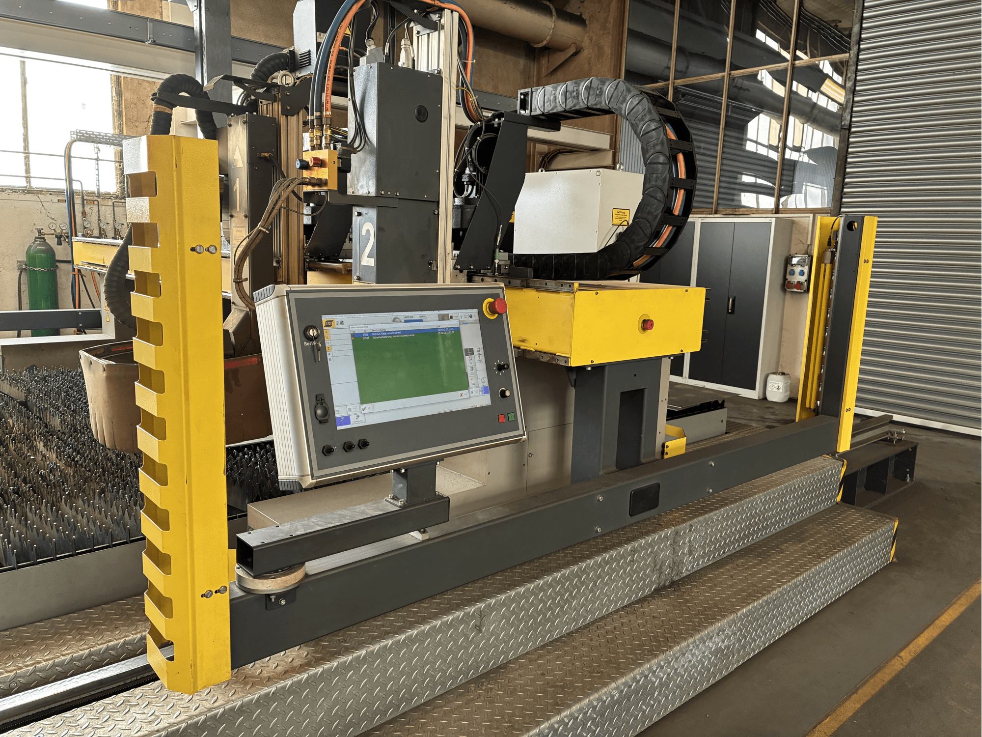 Industriële snijmachine met een touchscreen bedieningspaneel, gele veiligheidsafscherming, vooraanzicht, zichtbare metalen roosterbasis.