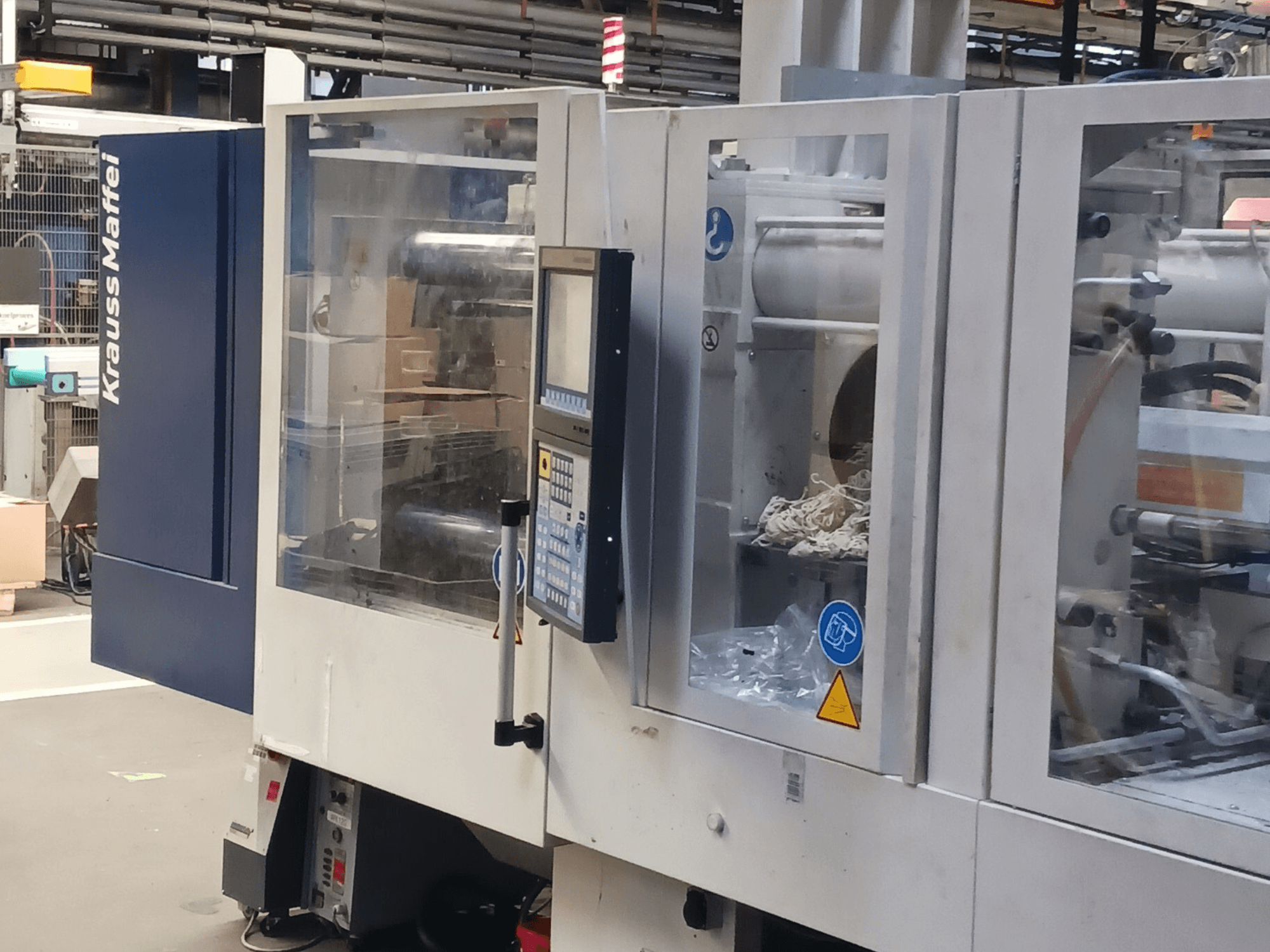 KraussMaffei spuitgietmachine, zijaanzicht, met bedieningspaneel, machines en verpakkingsmateriaal op de achtergrond.