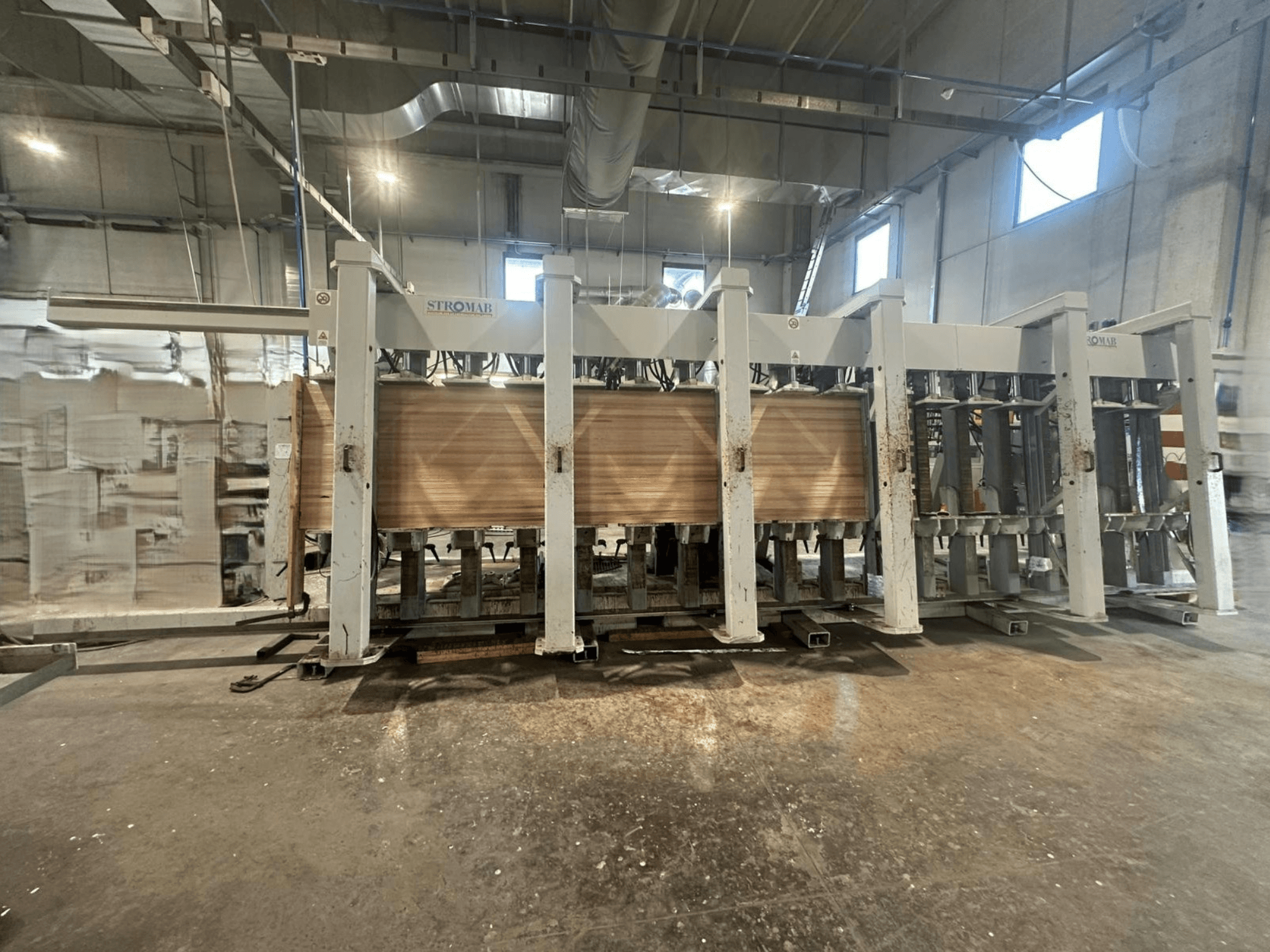 Industriële STROMAB machine in een magazijn, vooraanzicht, met houten panelen die worden bewerkt, met de metalen structuur en mechanische onderdelen.
