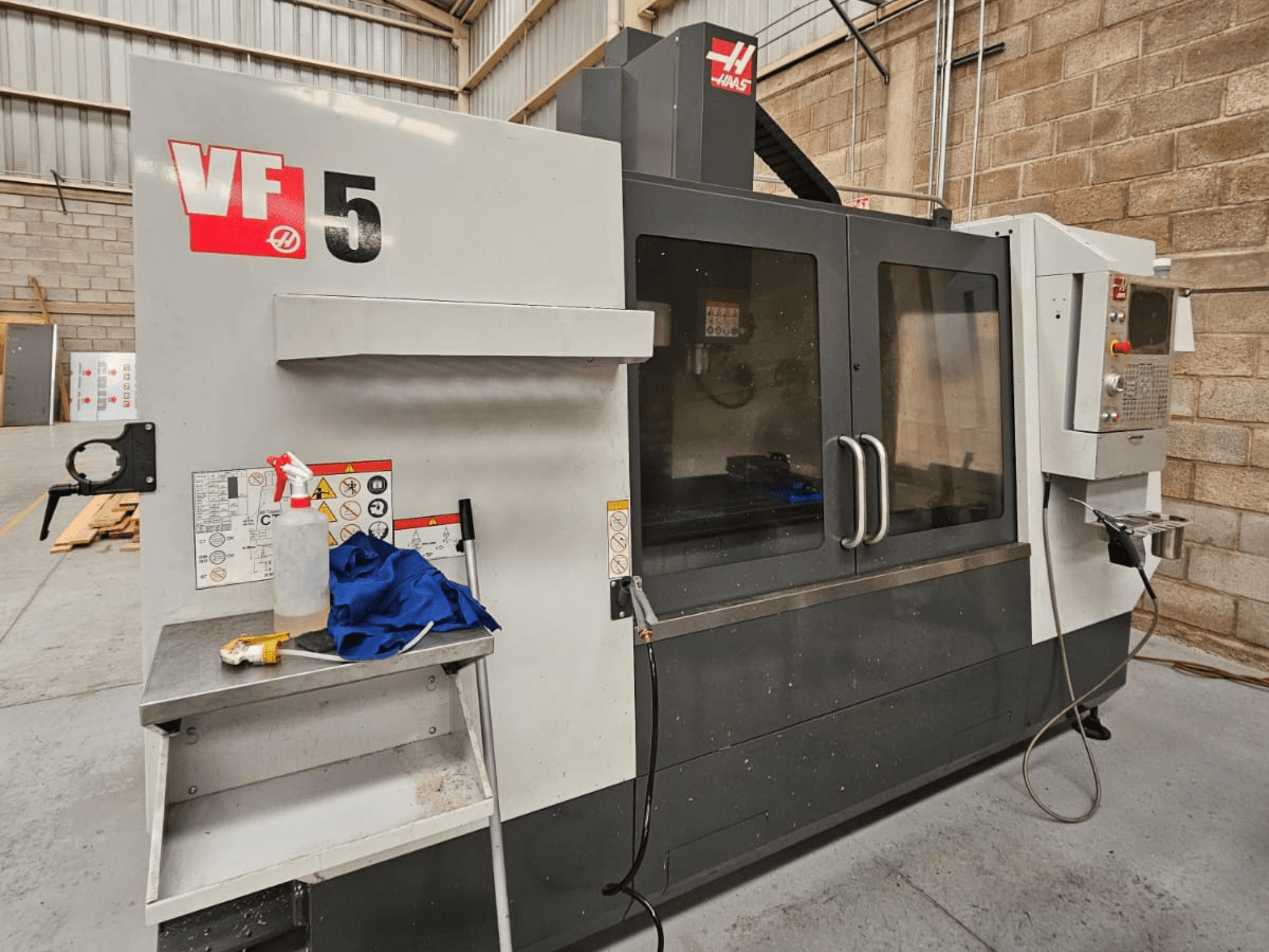Haas VF-5 CNC freesmachine in vooraanzicht, met bedieningspaneel, fles koelvloeistof, blauwe doek en veiligheidsinstructies.
