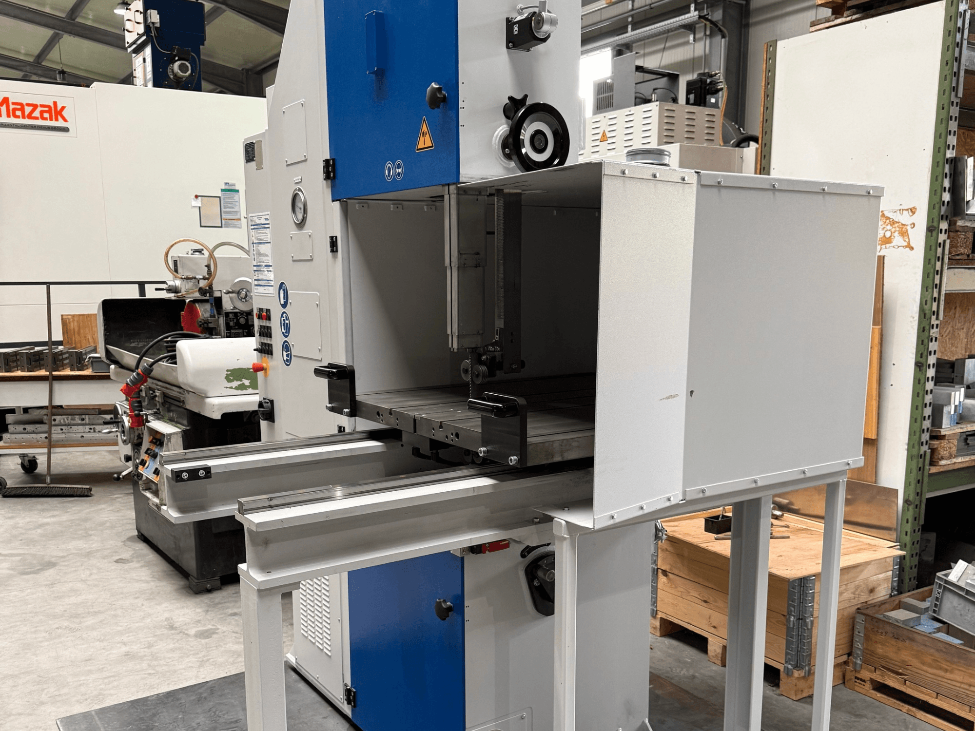 Mazak industriële machine gezien vanaf de voorkant, met blauw bedieningspaneel, metalen tafel en veiligheidswaarschuwingen.