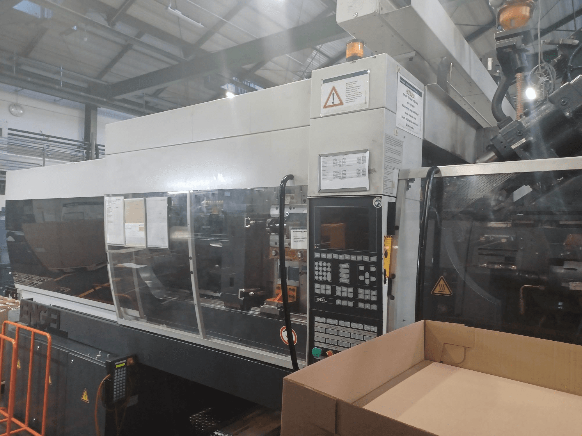 Engel spuitgietmachine, vooraanzicht, met bedieningspaneel, veiligheidslabels en een lege doos in de buurt.