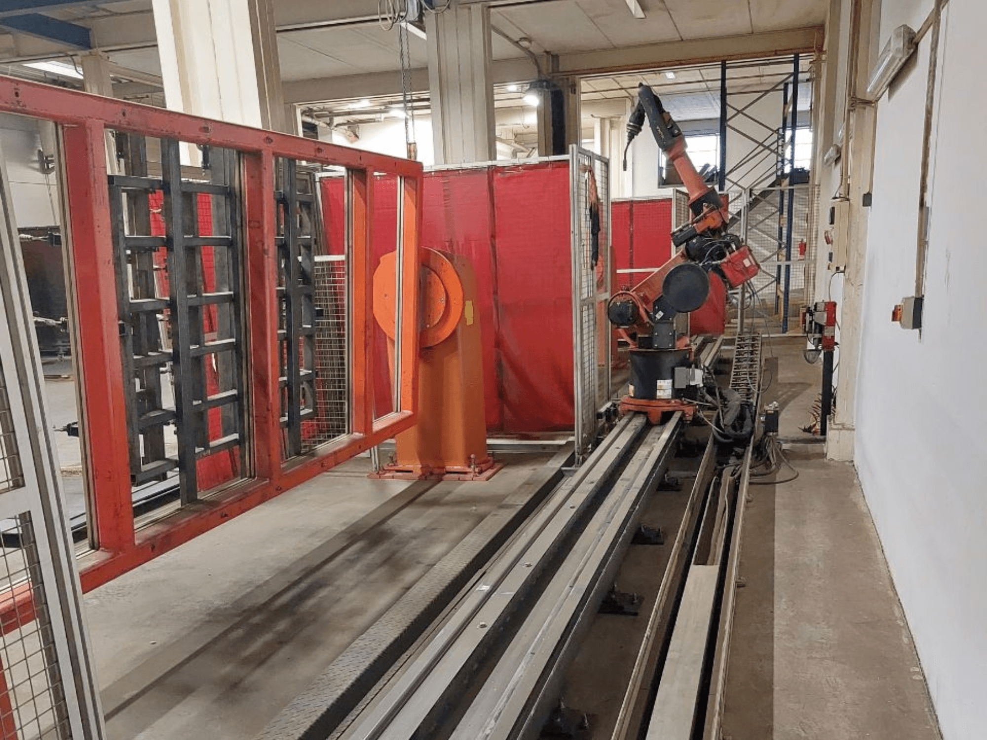 Industriële robotarm met oranje en zwarte kleuren op metalen rupsbanden, omgeven door rode beschermende barrières, vooraanzicht.