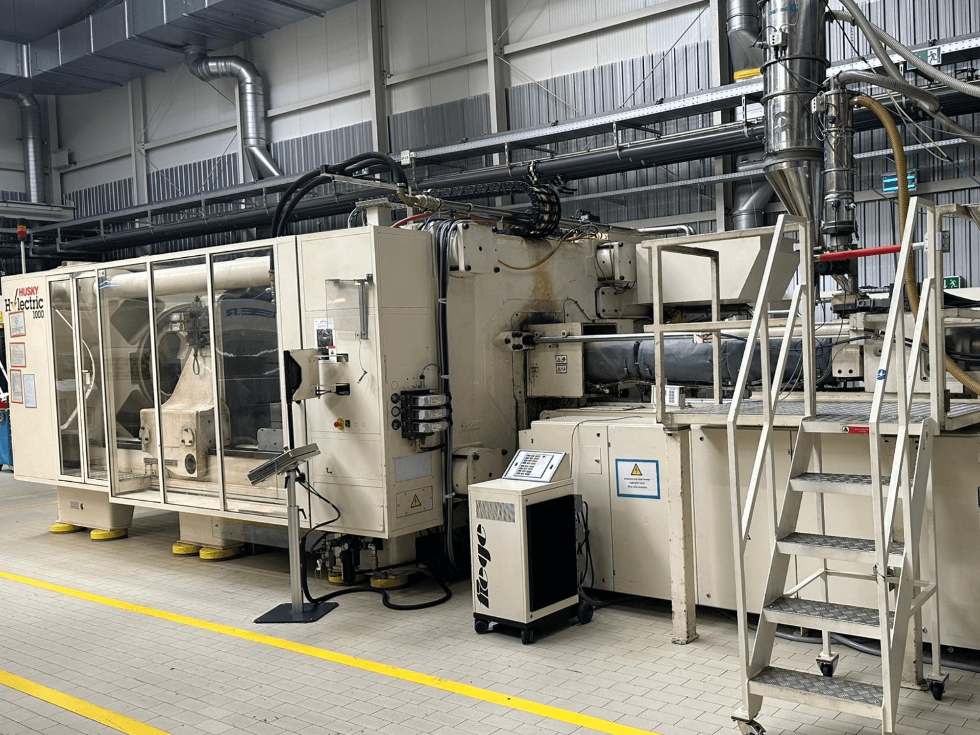 Vooraanzicht van de Husky Hylelectric H1000 RS95/80 elektrische spuitgietmachine in een fabrieksomgeving, met bedieningspaneel en traptreden.