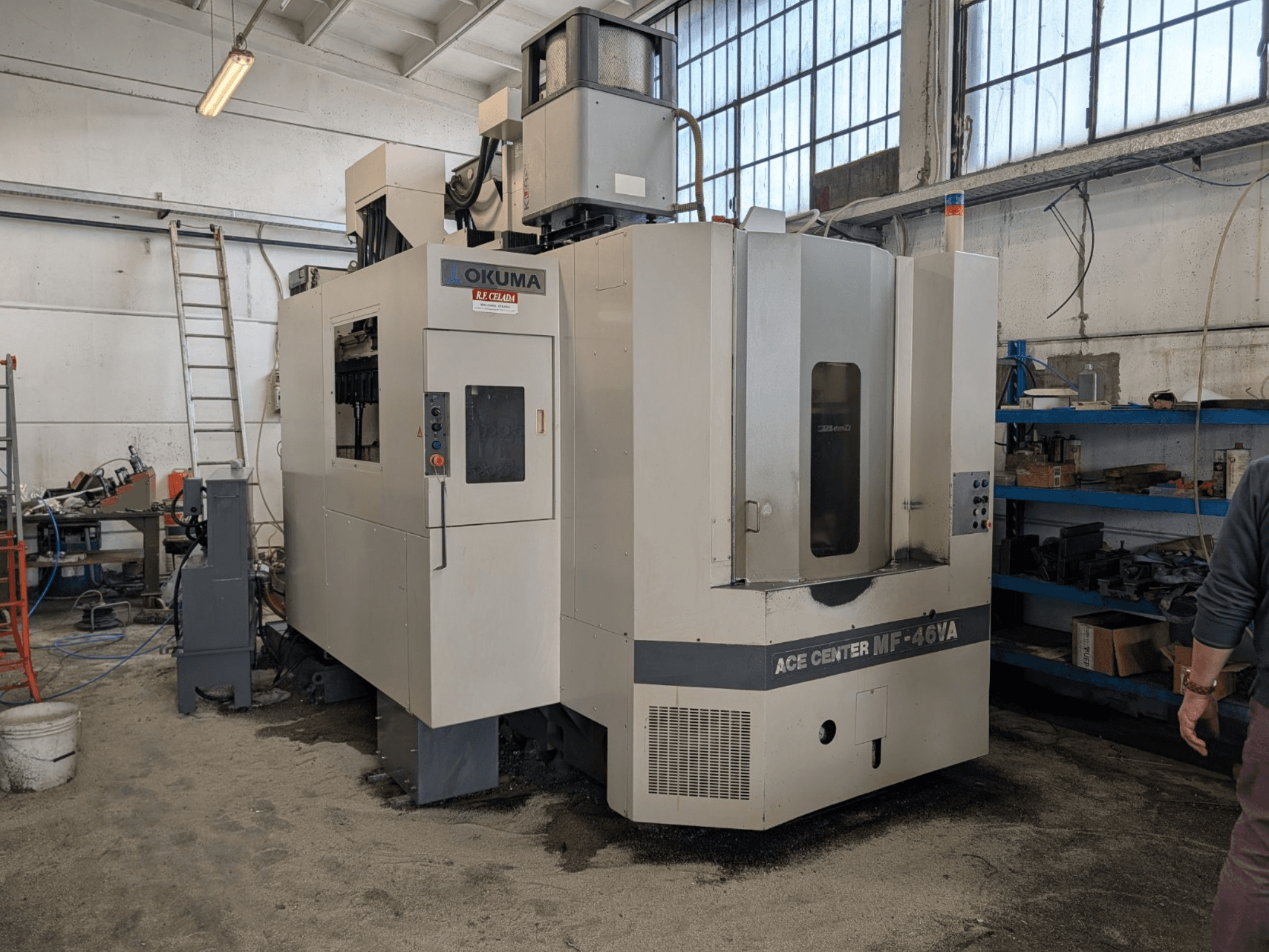 Okuma ACE CENTER MF-46VA verticaal bewerkingscentrum in een werkplaats, vooraanzicht met bedieningspaneel en omringende apparatuur.