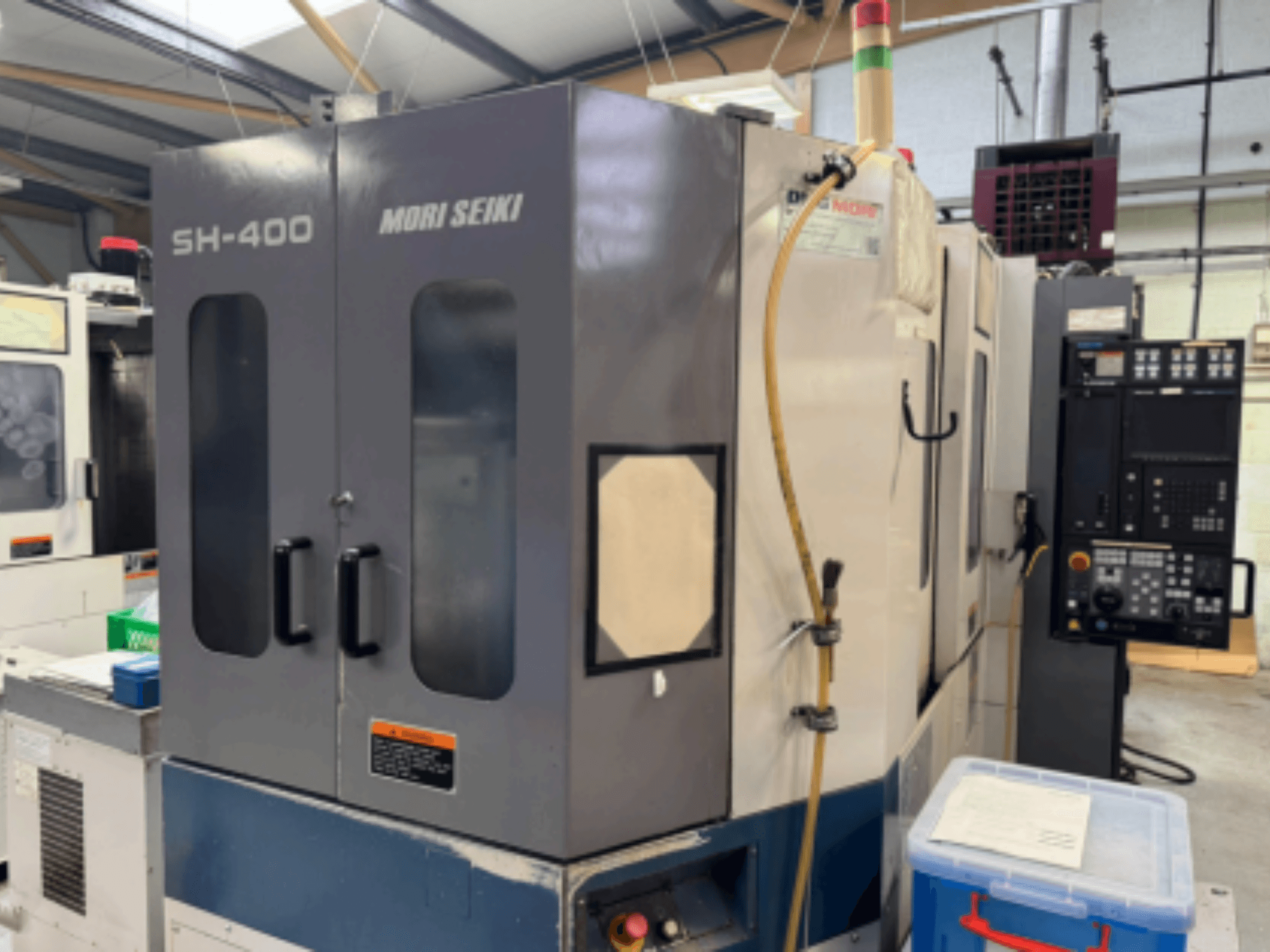 Mori Seiki SH-400 CNC machine, vooraanzicht, met dubbele deuren, bedieningspaneel en bevestigingsslang. Blauwe opslagcontainer vlakbij.