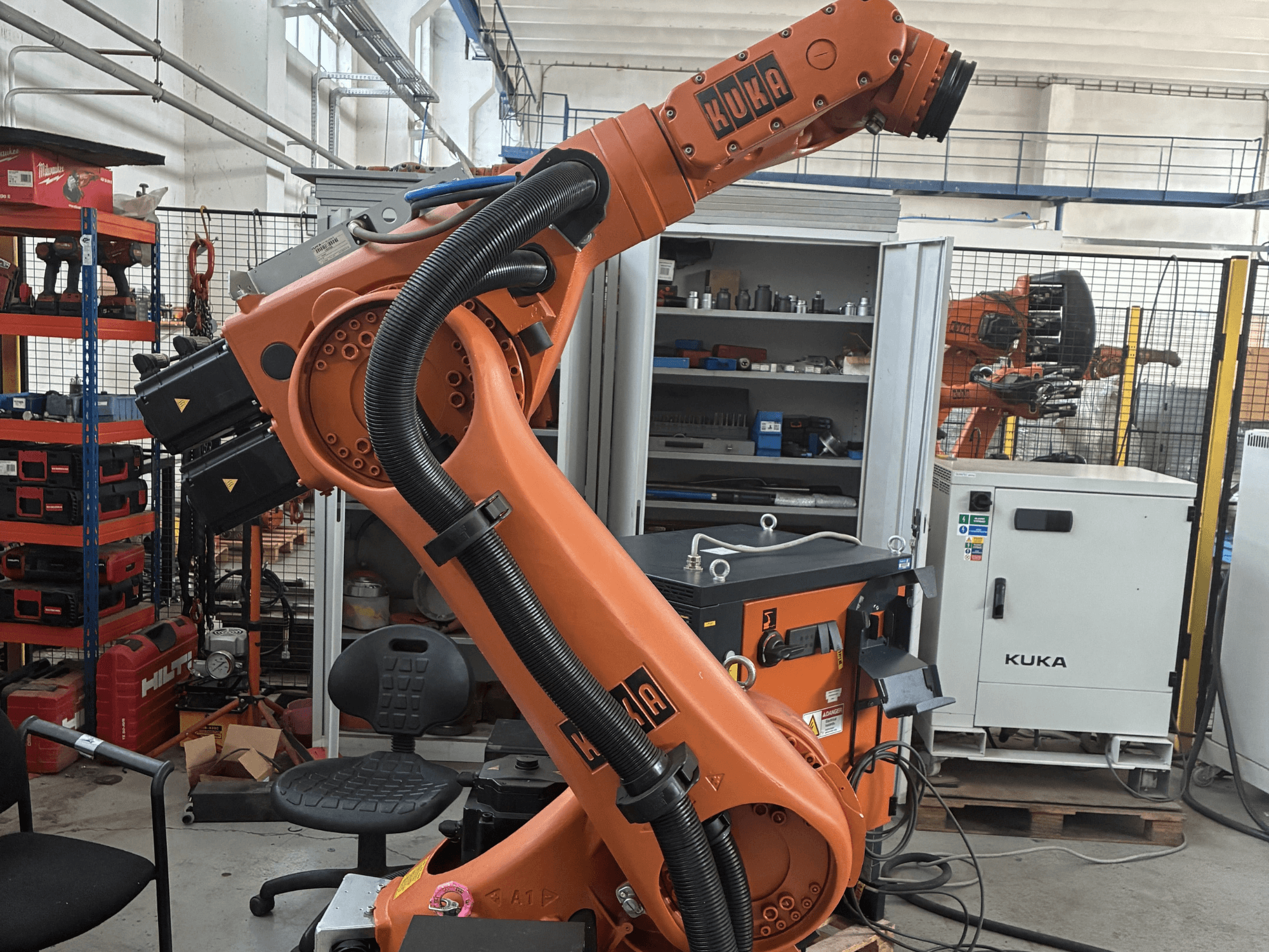 KUKA KR30-3 robotarm in een industriële omgeving, zijaanzicht met kabels en bedieningselementen, met gereedschap en rekken op de achtergrond.
