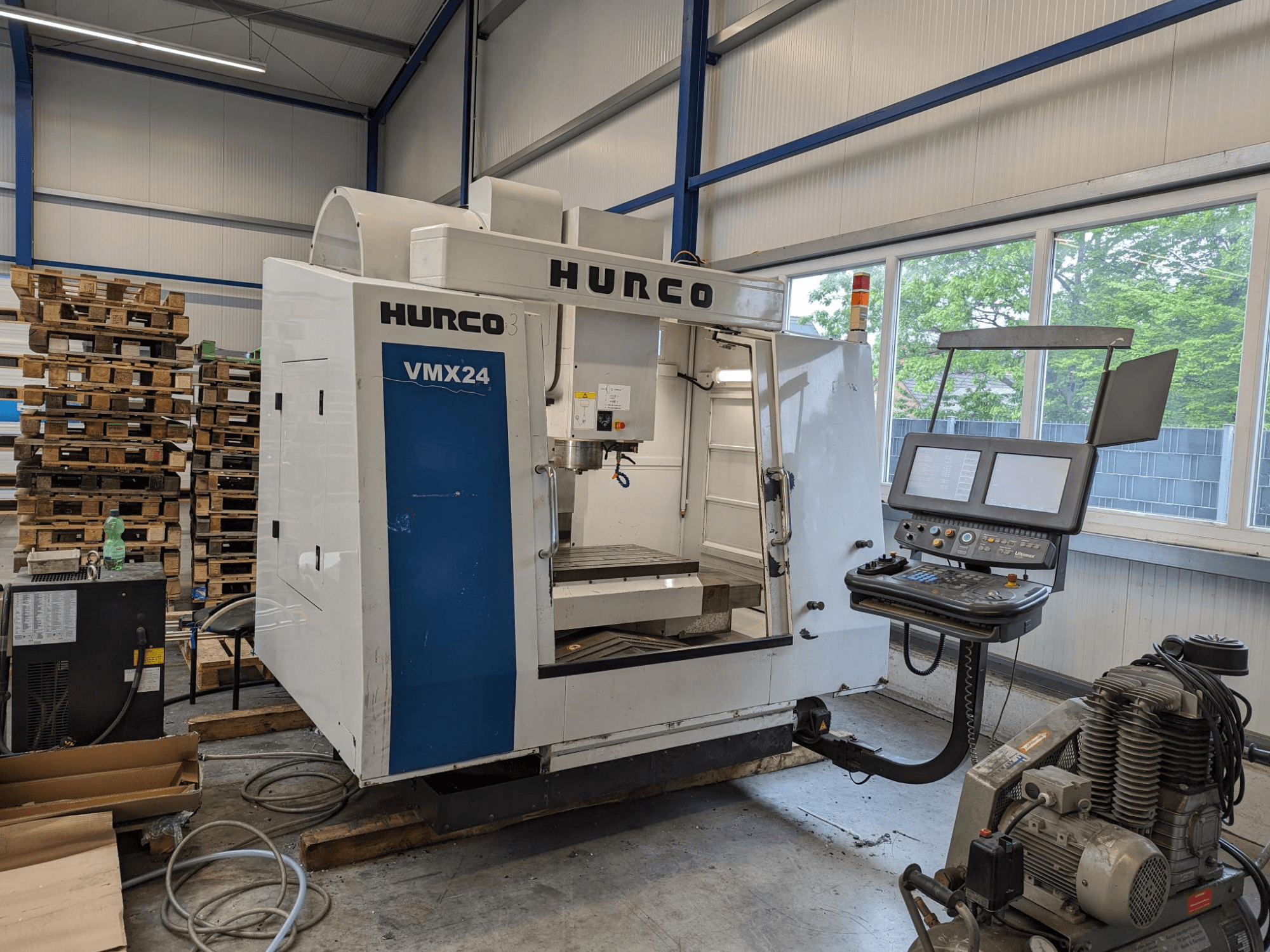 Hurco VMX24 freesmachine, vooraanzicht, met bedieningspaneel en gereedschapgebied; gestapelde pallets op achtergrond; industriële werkruimte.
