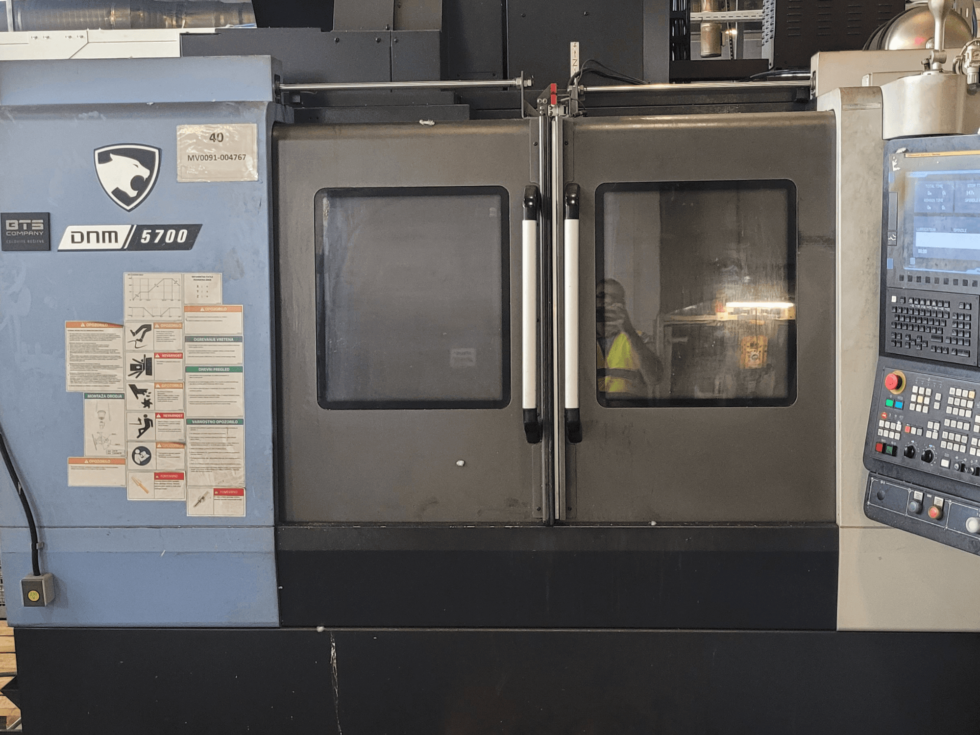 DOOSAN DNM 5700 verticaal bewerkingscentrum, vooraanzicht met bedieningspaneel, veiligheidsstickers en deuren die op een kier staan.
