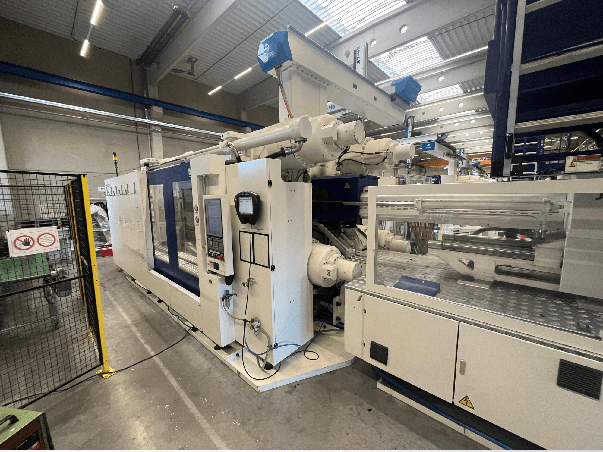 Spuitgietmachine in wit en blauw, vooraanzicht, met bedieningspaneel, robotarm en veiligheidsborden.