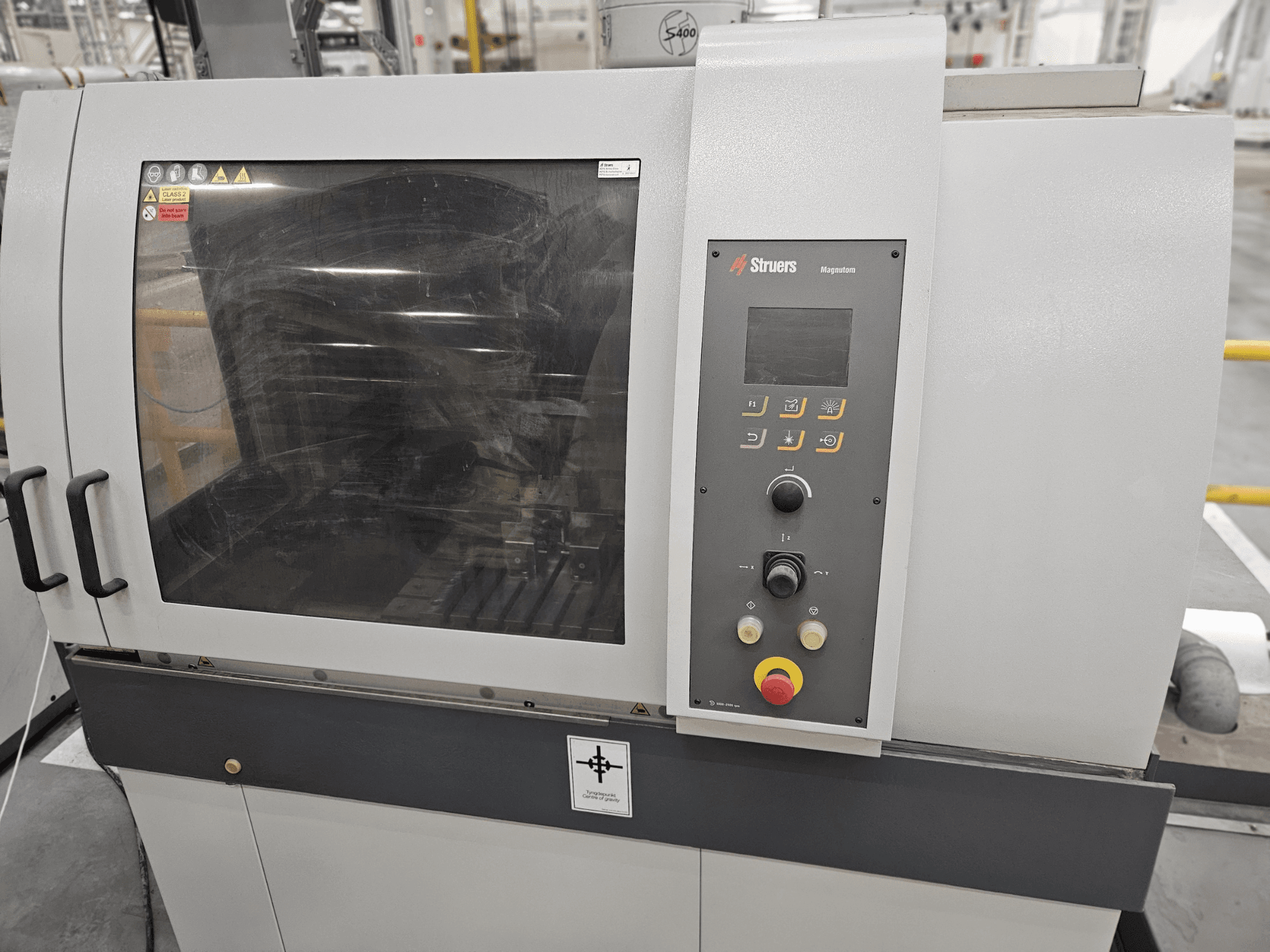 Struers Magpentor machine vooraanzicht met bedieningspaneel, knoppen en een groot kijkvenster. Technische labels zichtbaar.