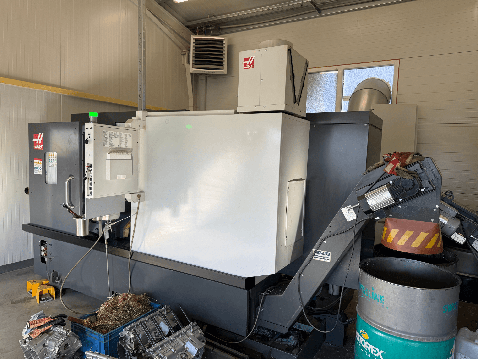 Haas CNC machine met bedieningspaneel, zijaanzicht; zichtbare koelvloeistof- en olieleidingen; metaalschroot in blauwe bak; industriële omgeving.