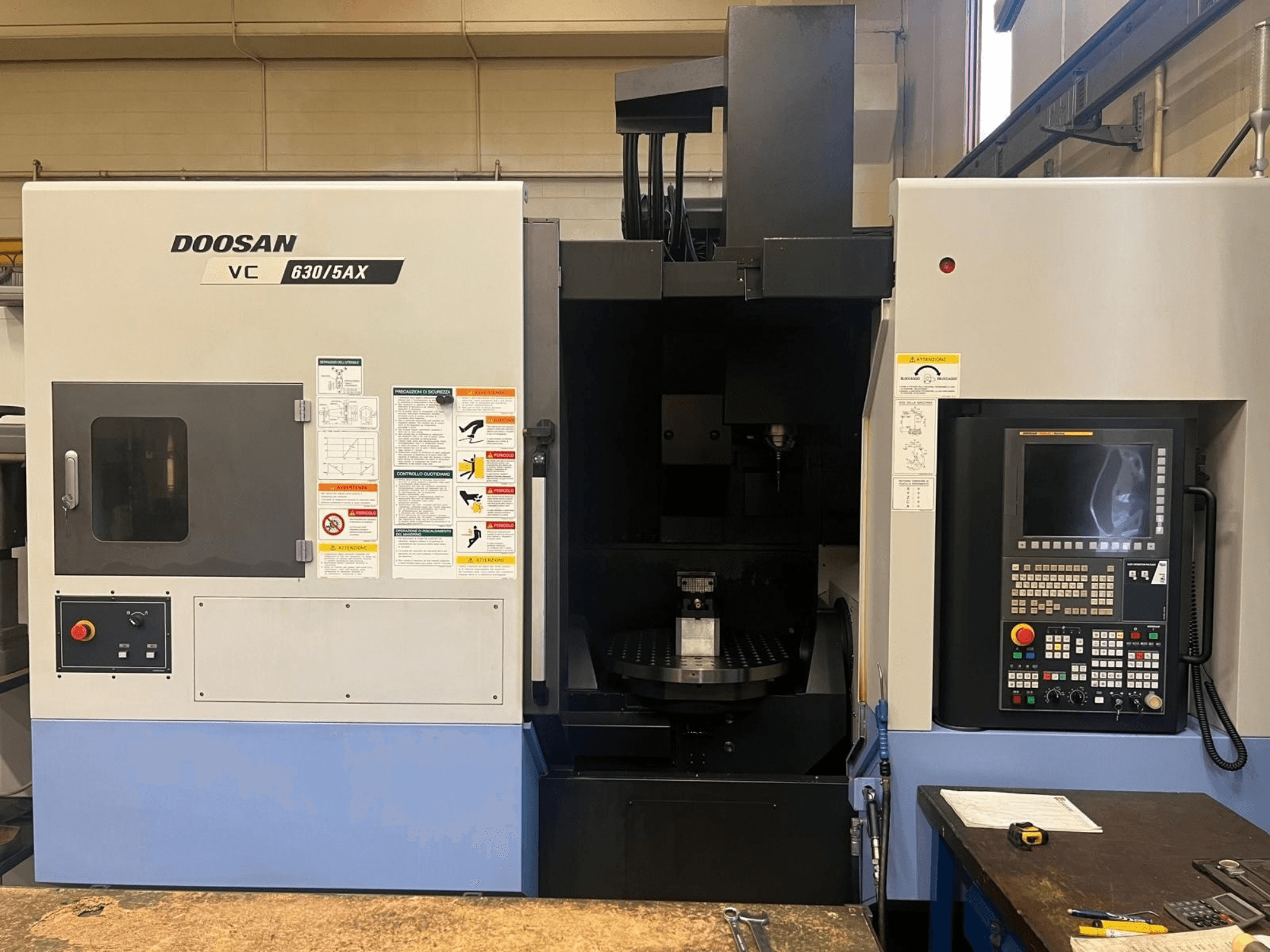 Vooraanzicht van een DOOSAN VC 630/5AX verticaal bewerkingscentrum, met het bedieningspaneel en het spilgedeelte in beeld, inclusief de zichtbare veiligheidsstickers.